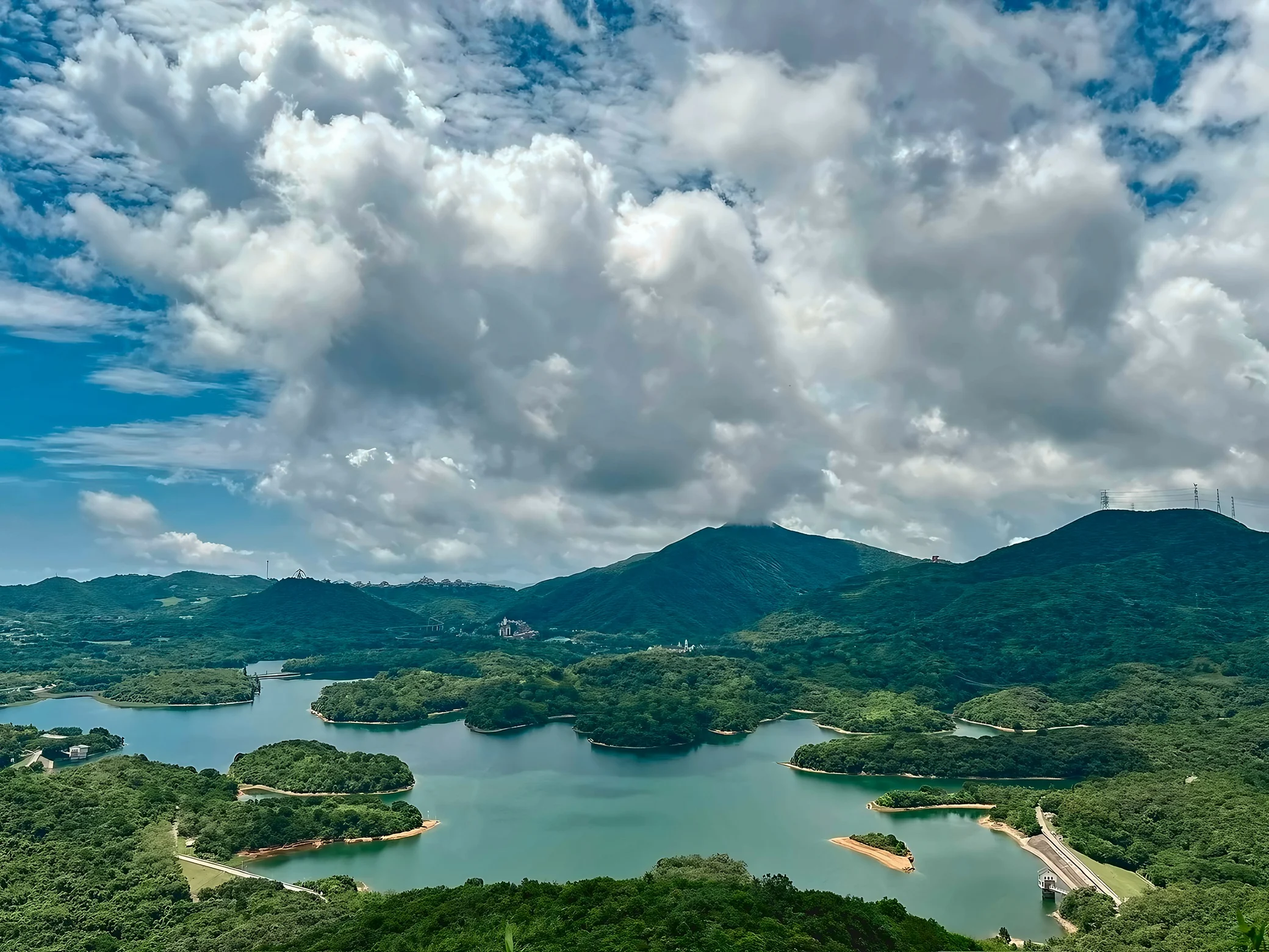 A winding turquoise reservoir lies among forested hills beneath large white clouds, with a road and waterworks along the right bank and transmission towers on a ridge.