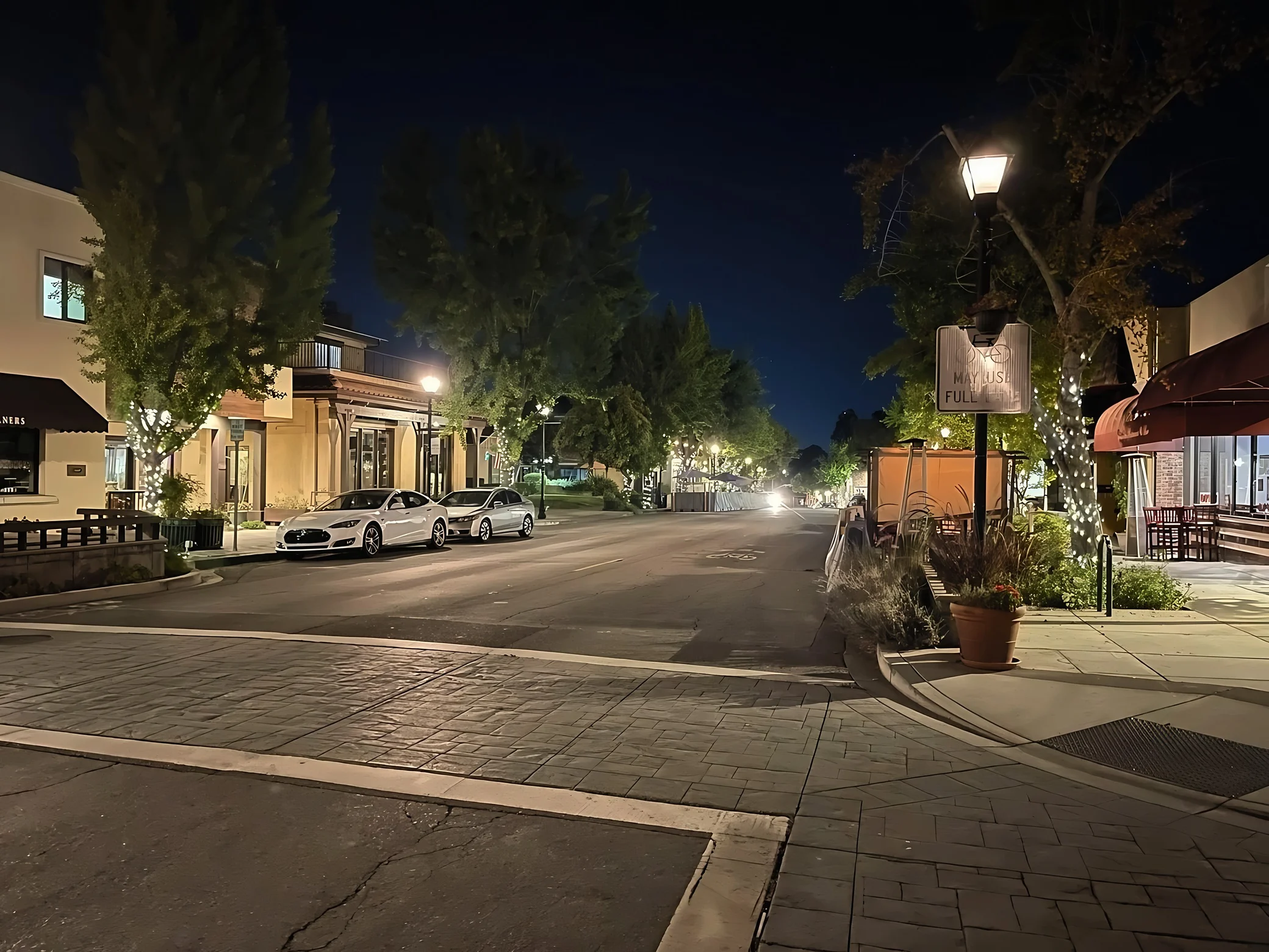 A small-town California street is lined with shops, illuminated trees and parked cars beside a bicycle sign reading “MAY USE FULL LANE.”