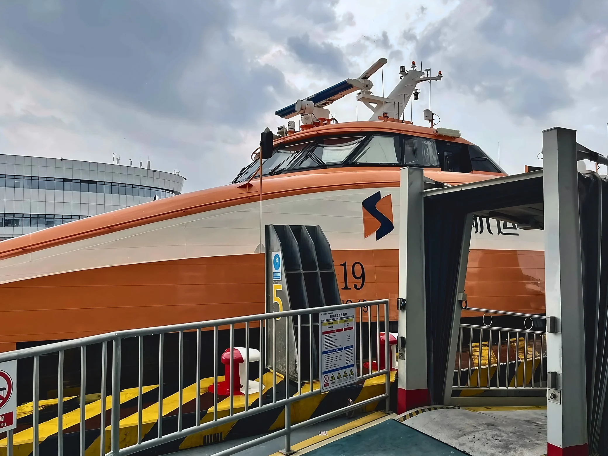 An orange-and-white high-speed ferry is moored beside a boarding gangway, metal railings, and yellow-and-black dock markings under a cloudy sky.