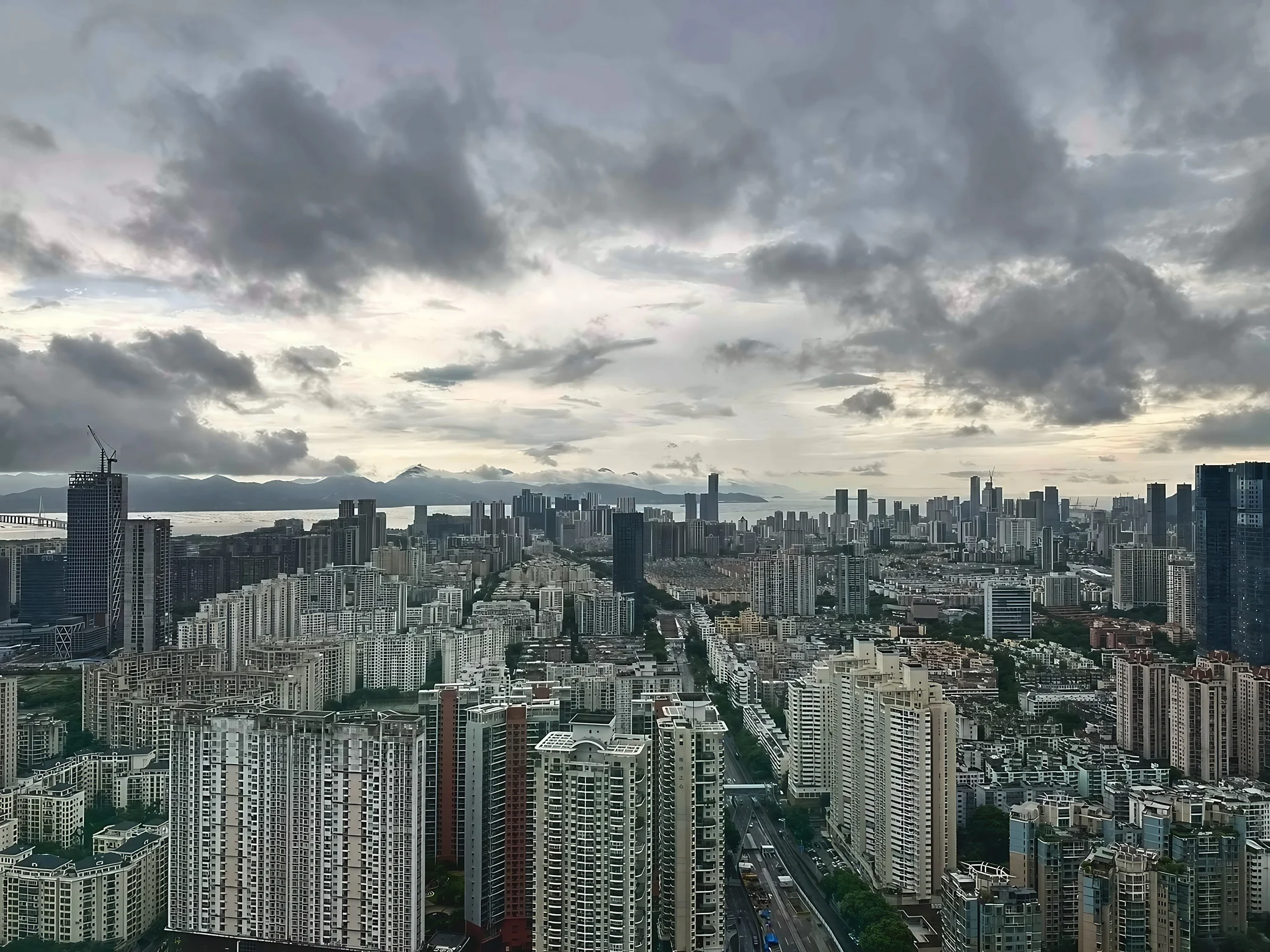Dense residential towers and office buildings stretch toward a bay and distant mountains under a cloudy sky in Shenzhen.
