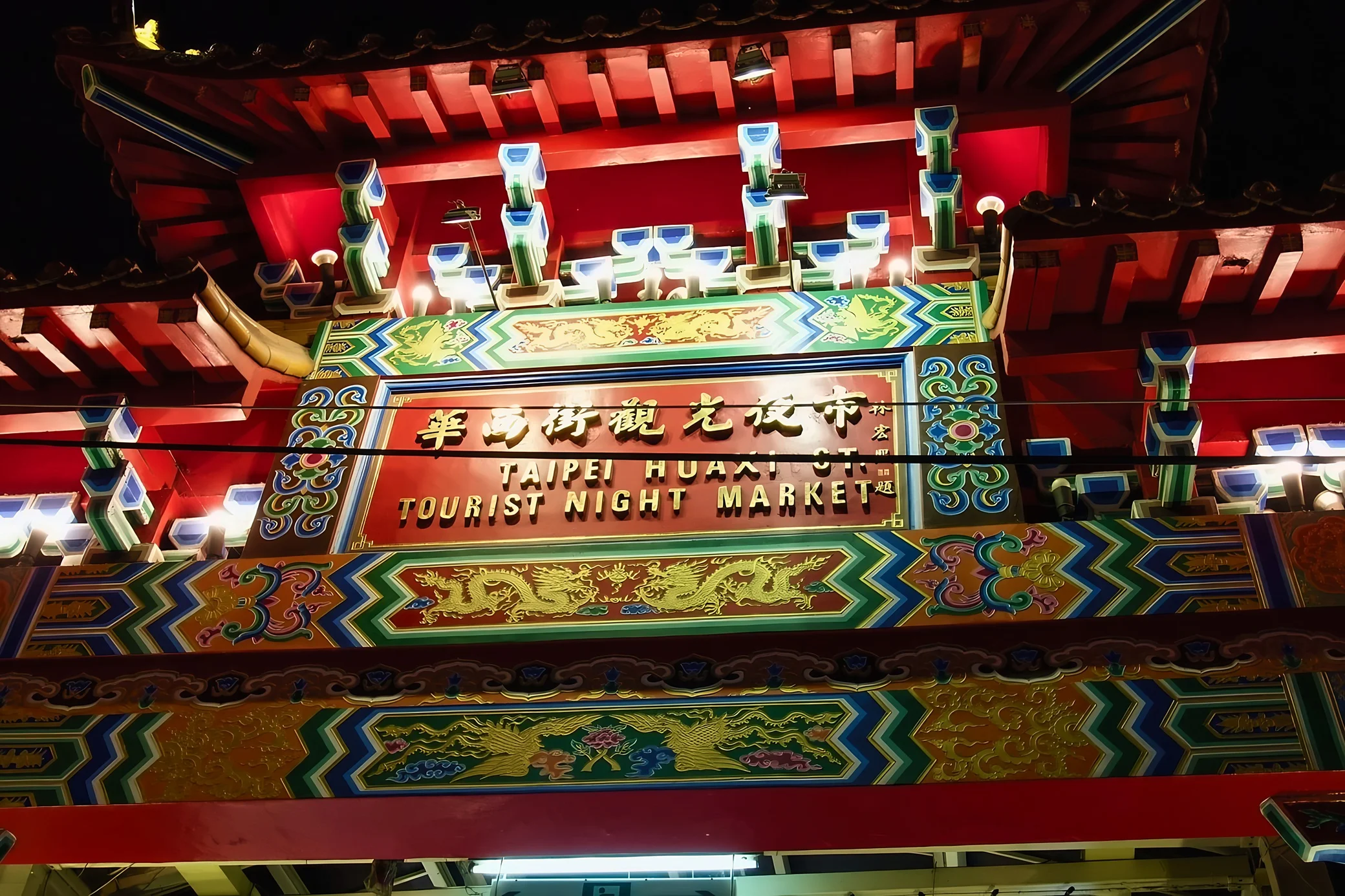 The red Huaxi Street Tourist Night Market gateway features a bilingual sign, golden dragon motifs and brightly painted beams crossed by overhead wires.