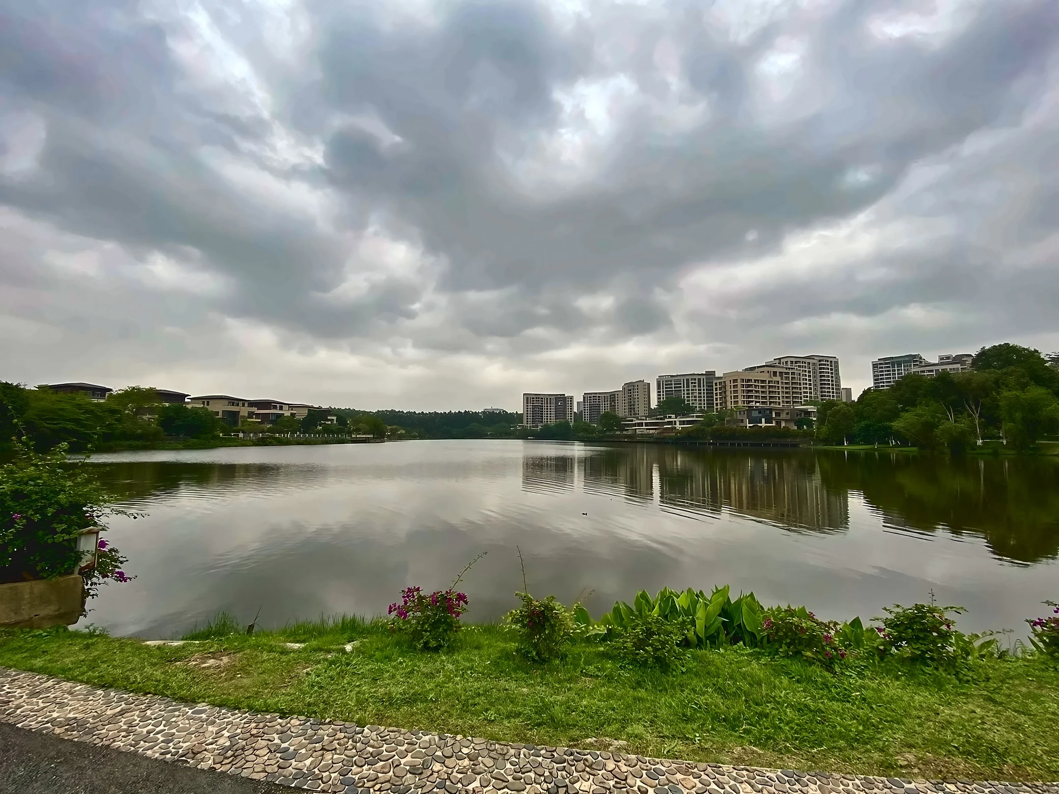 A wide view of the lake shows cloudy reflections, waterfront buildings and trees beyond a foreground of grass, purple flowers and a pebble-edged path.