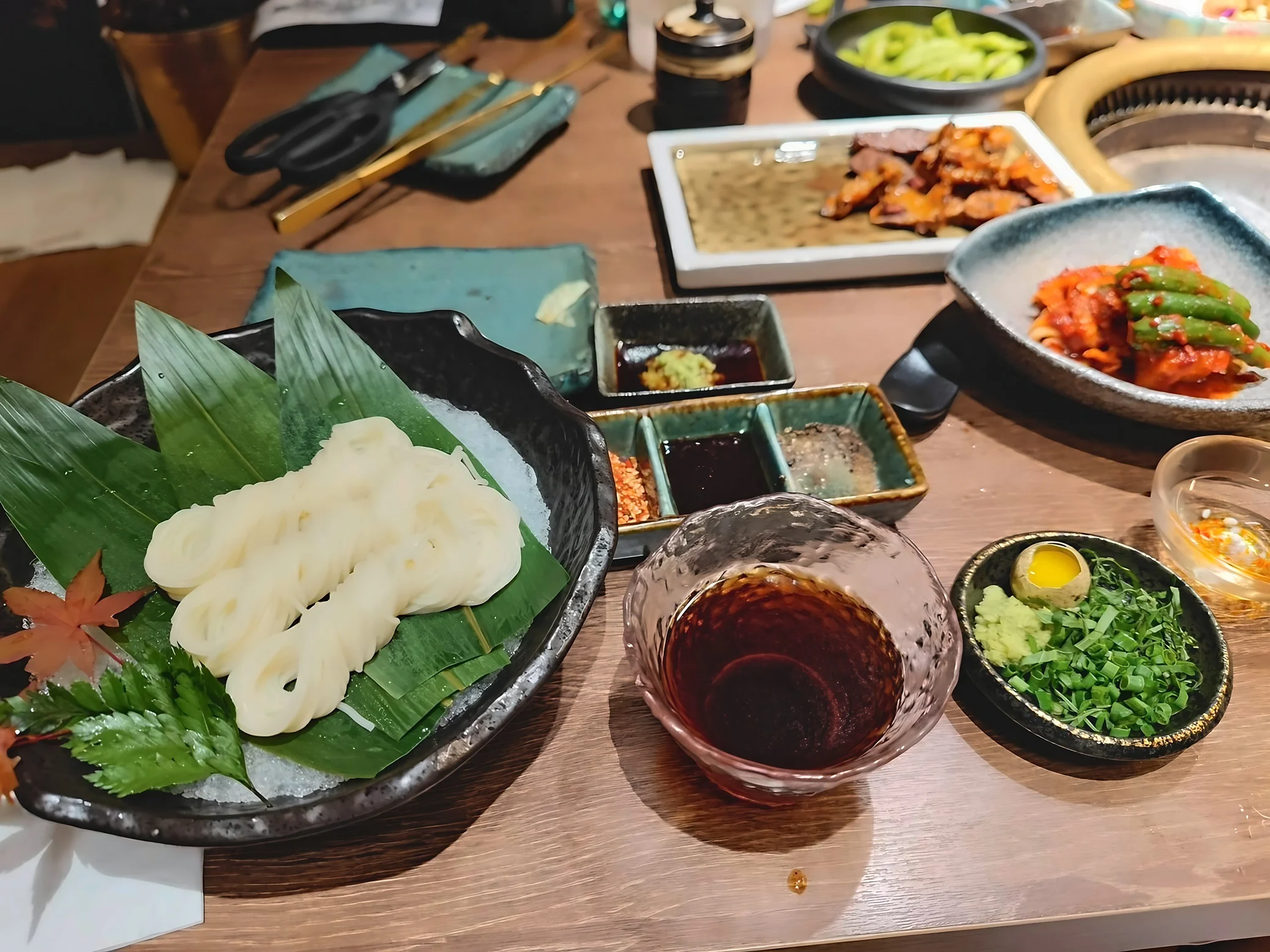 Chilled white noodles rest on green leaves over ice beside dipping sauce, scallions, a quail egg yolk, kimchi and sliced meat at a tabletop grill.