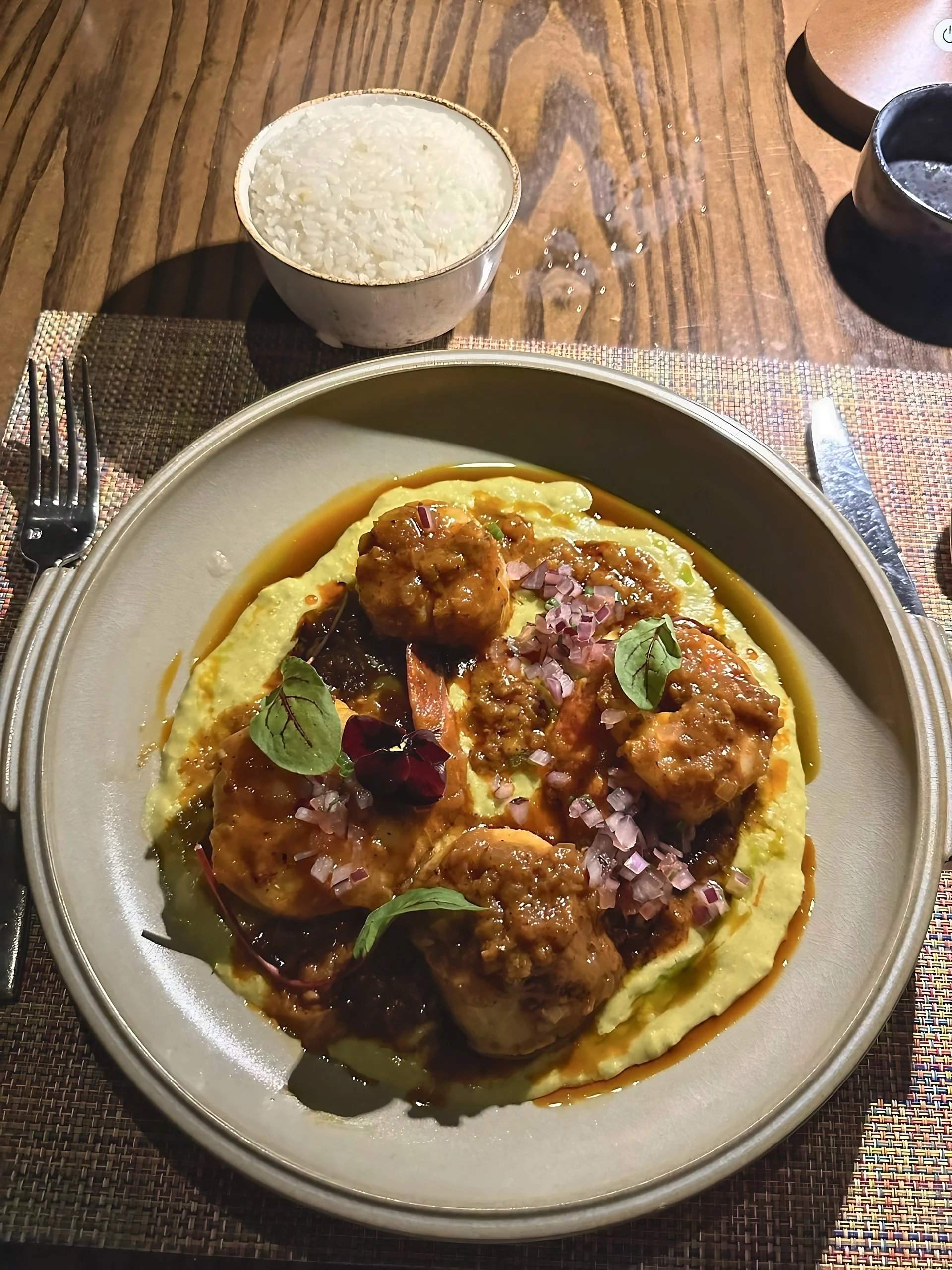 A close-up shows shrimp in a reddish-brown sauce over a yellow base, garnished with diced onion, leaves and a purple flower, with rice above the plate.