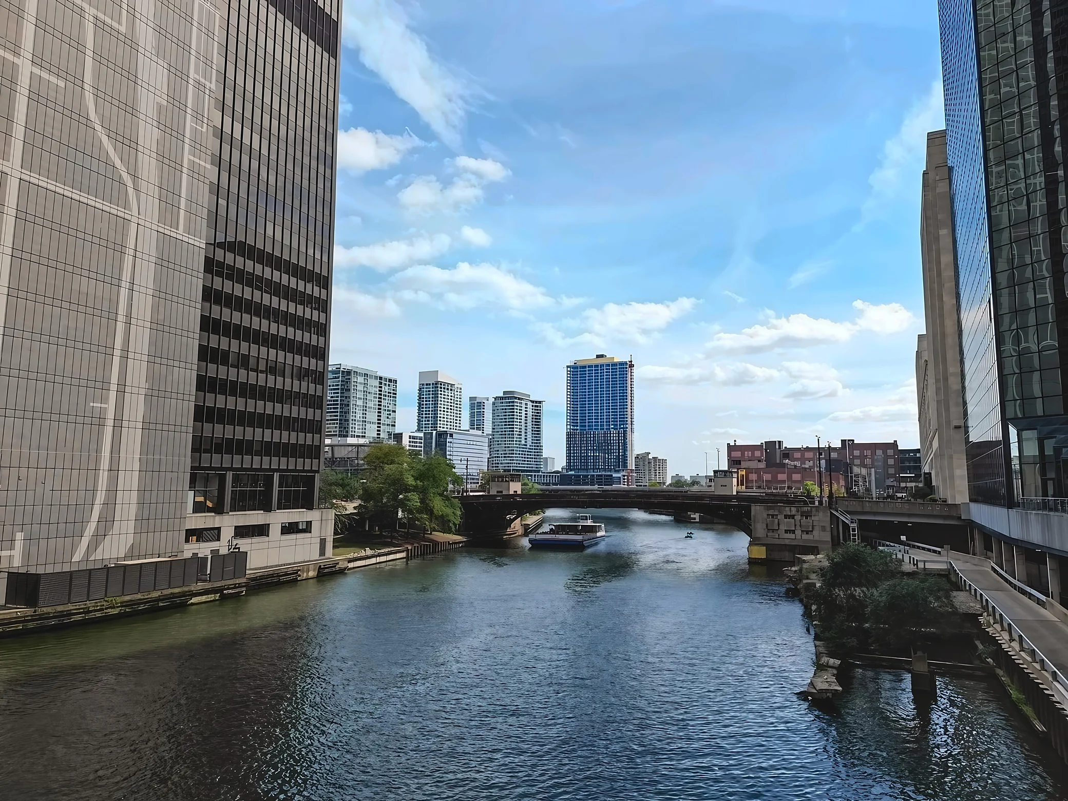 Under a bright blue sky, the Chicago River runs between tall buildings toward a steel bridge, with a white tour boat and a small green craft on the water.