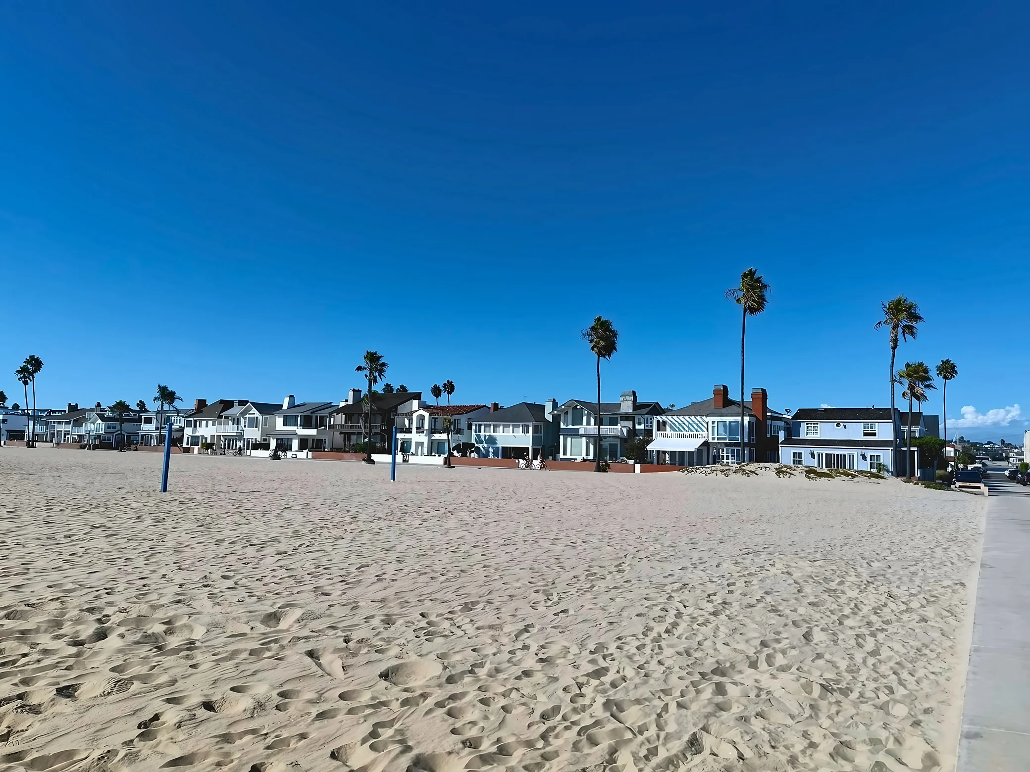 A row of beachfront homes stands beneath tall palm trees, with footprints and tire tracks in the foreground sand and a concrete path leading to the street on the right.