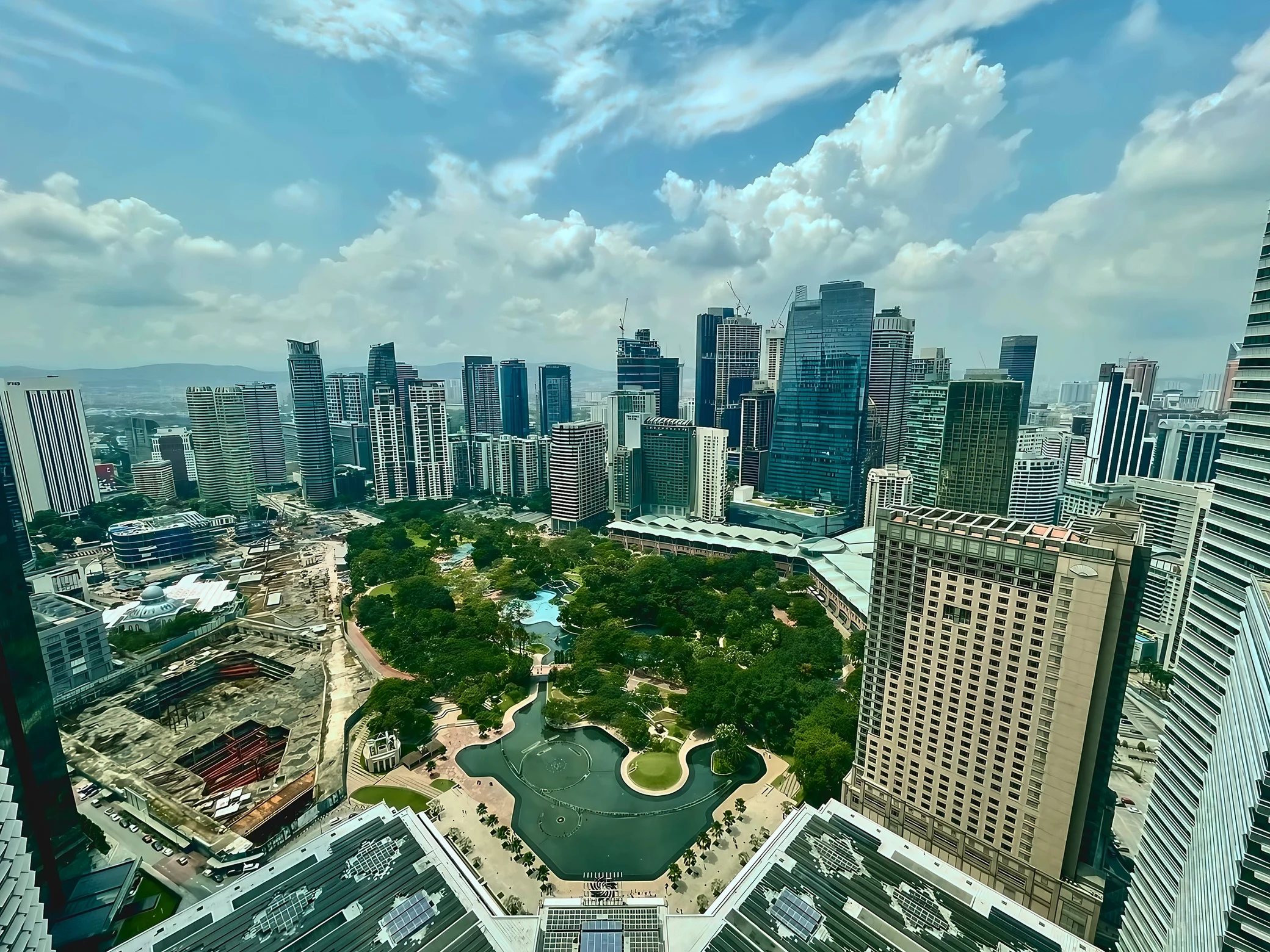 Towers surround KLCC Park and its fountain lake beneath a cloud-filled blue sky, with a construction site on the left and hotel buildings on the right.