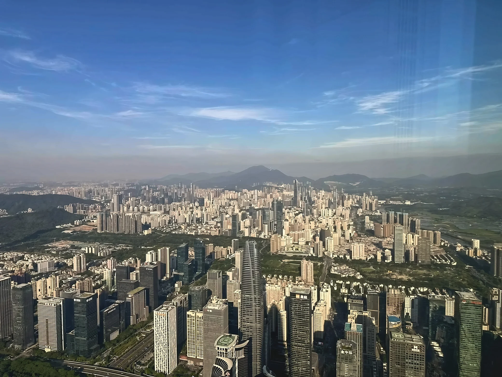 Shenzhen's towers, green spaces and roads extend toward distant mountains beneath a blue sky streaked with thin clouds.