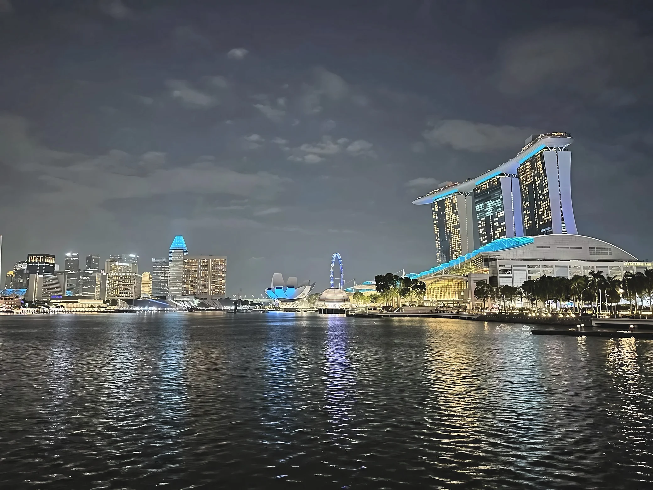 Marina Bay Sands towers above the waterfront beside the lotus-shaped ArtScience Museum and Singapore Flyer, with colorful reflections across the bay.