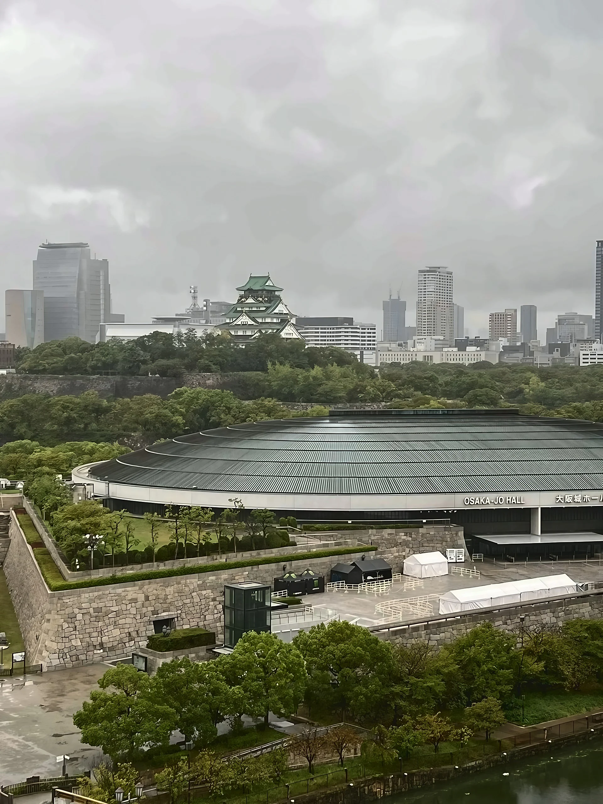 Osaka Castle rises beyond the trees behind Osaka-jo Hall, with stone walls, greenery, and a river in the foreground.