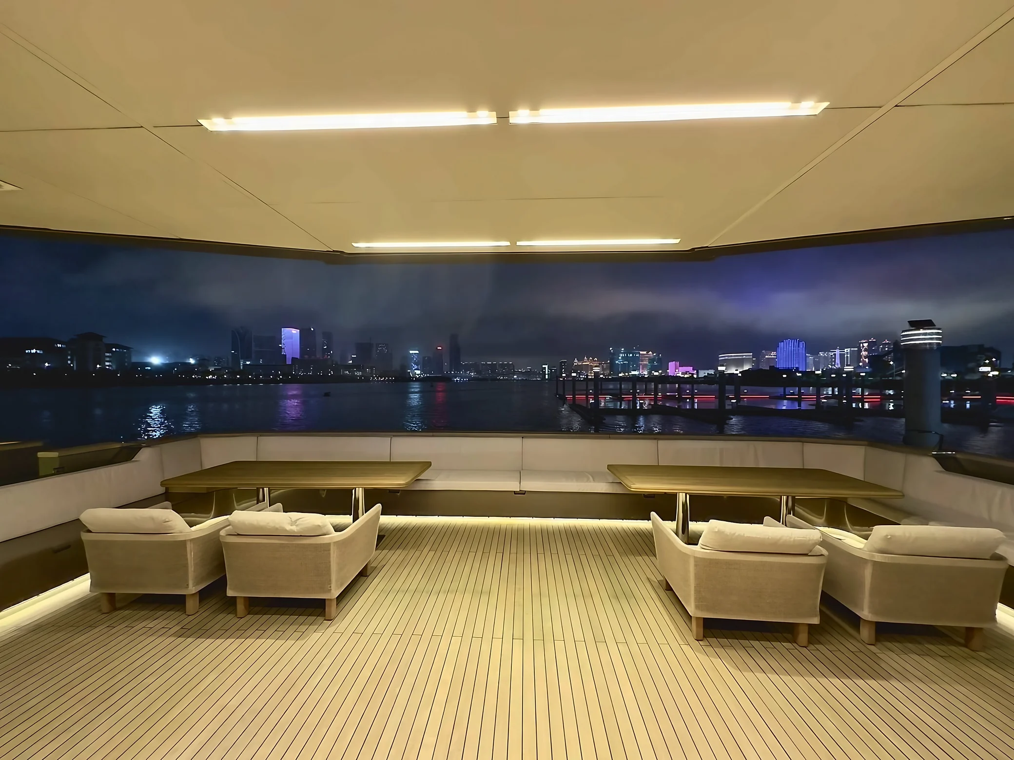 Four armchairs, two wooden tables and white bench seating occupy a covered yacht deck overlooking illuminated buildings across the water.
