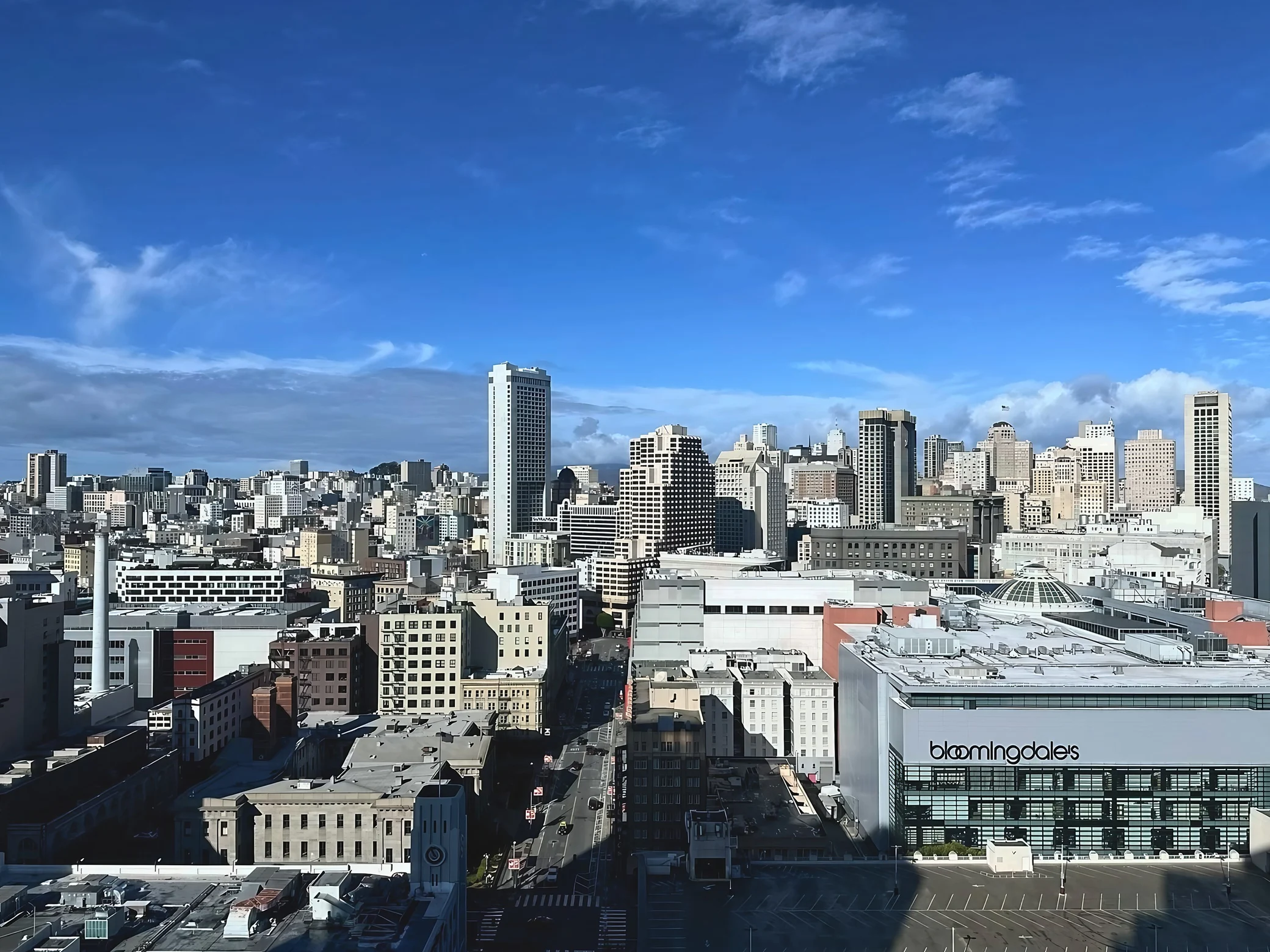 Downtown San Francisco's skyline rises above a straight street beside the clearly marked Bloomingdale's building under a blue sky.