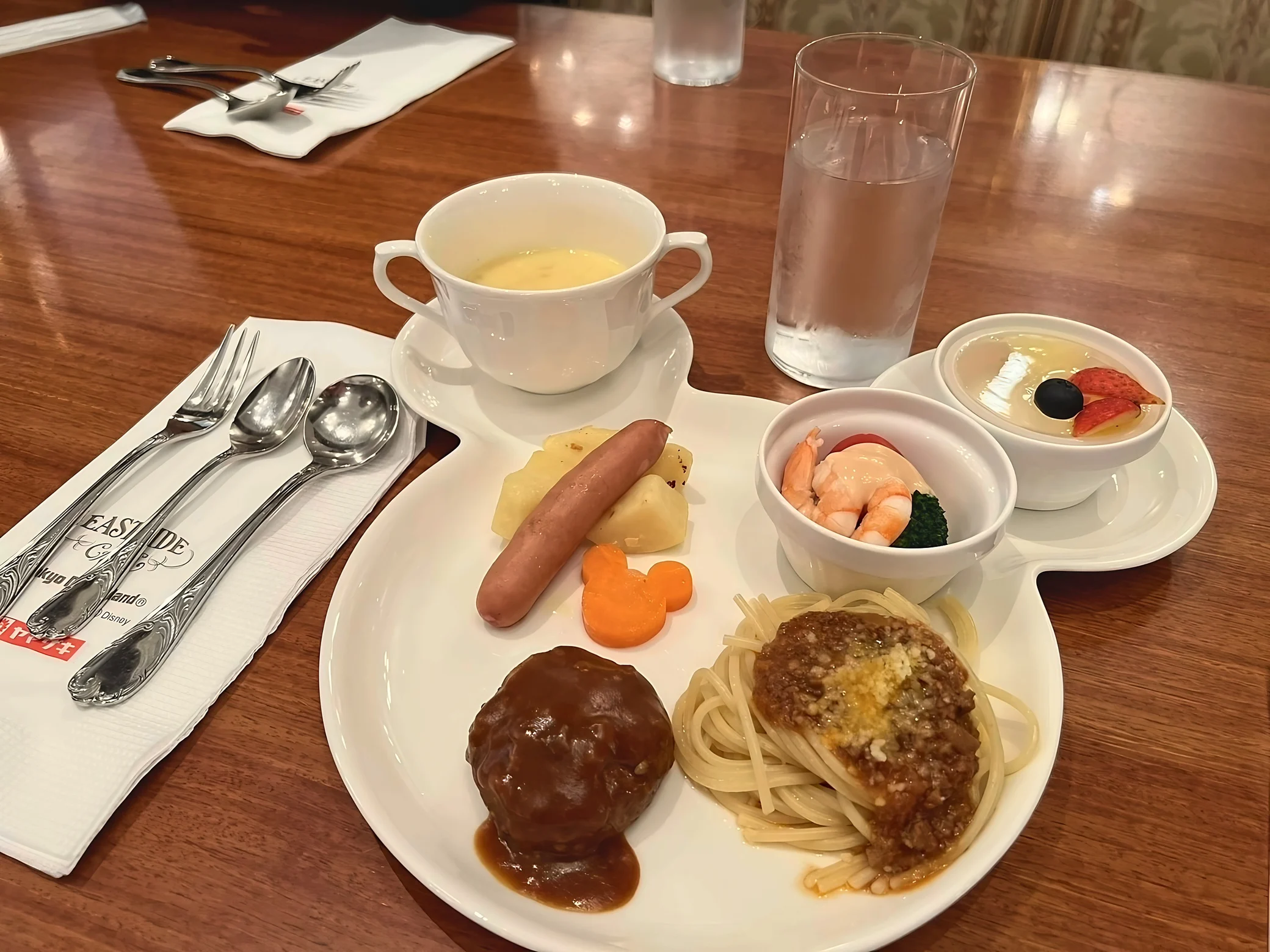 A close-up shows hamburg steak and spaghetti with meat sauce on a Mickey-shaped plate, with sausage, potatoes, a Mickey-shaped carrot, soup, shrimp salad and fruit dessert behind them.