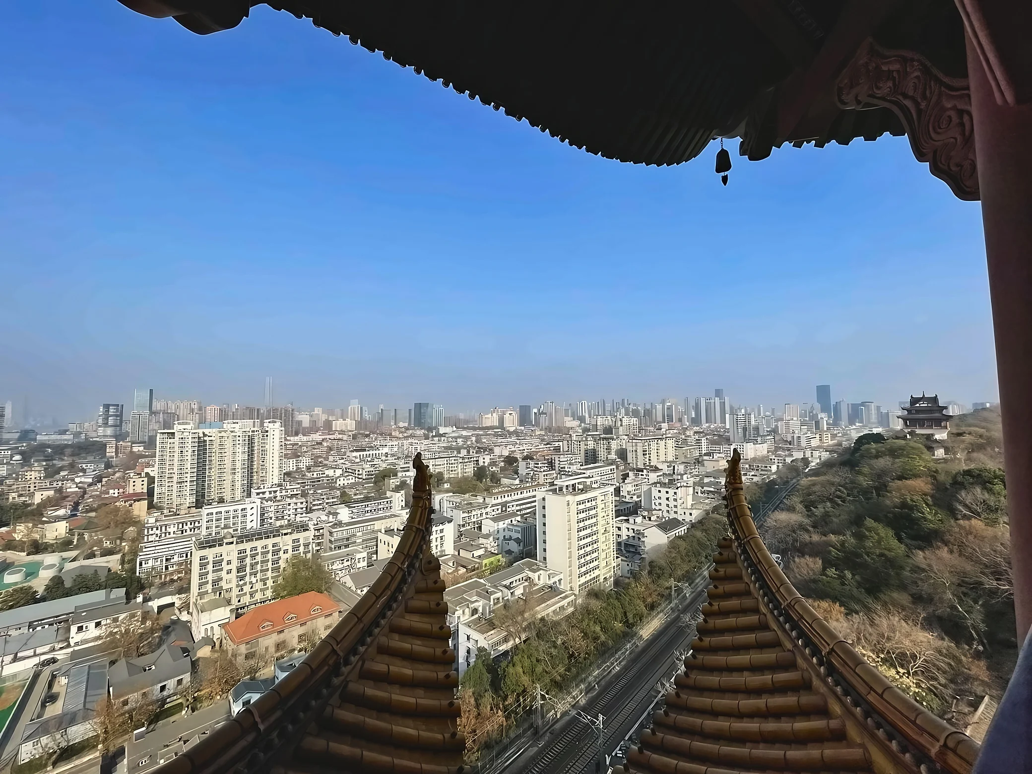 Traditional eaves and tiled roof ridges frame an elevated view of Wuhan, with railway tracks and a wooded hillside below.