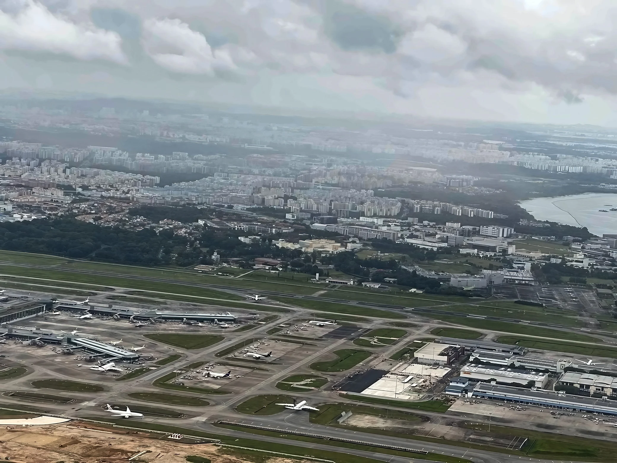 An expansive aerial view of Changi Airport shows terminal buildings, aprons, taxiways, runways, and numerous aircraft, with city neighborhoods and coastline beyond.
