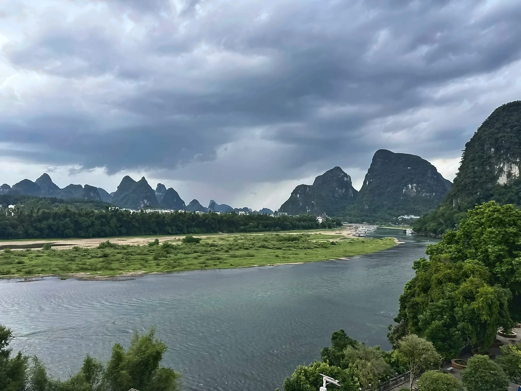 A river curves past a long green sandbar toward karst peaks and distant white boats beneath a dramatic canopy of gray clouds.