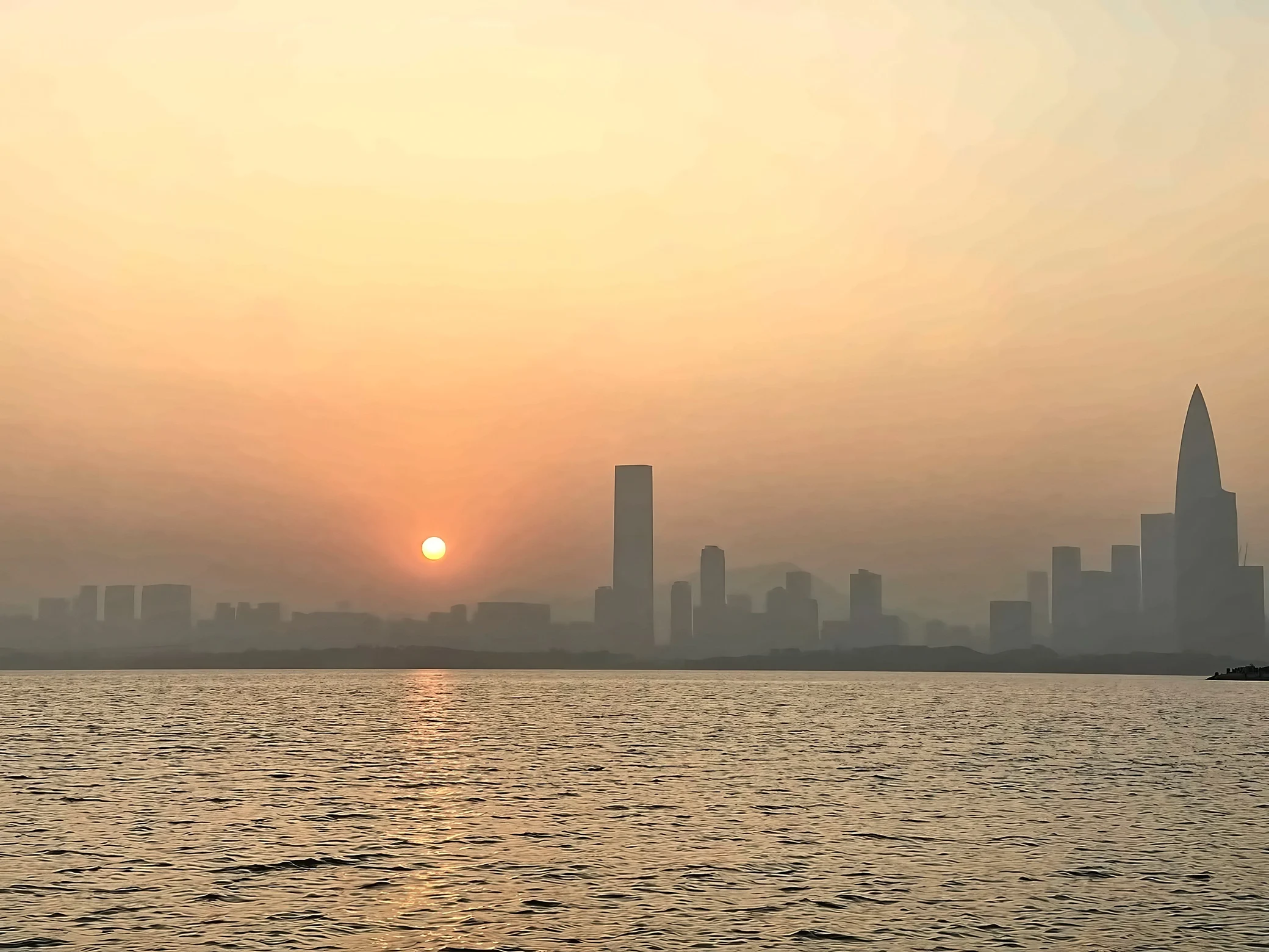 An orange sun casts a golden reflection across Shenzhen Bay beneath a hazy skyline, with the bamboo shoot-shaped China Resources Tower on the right.