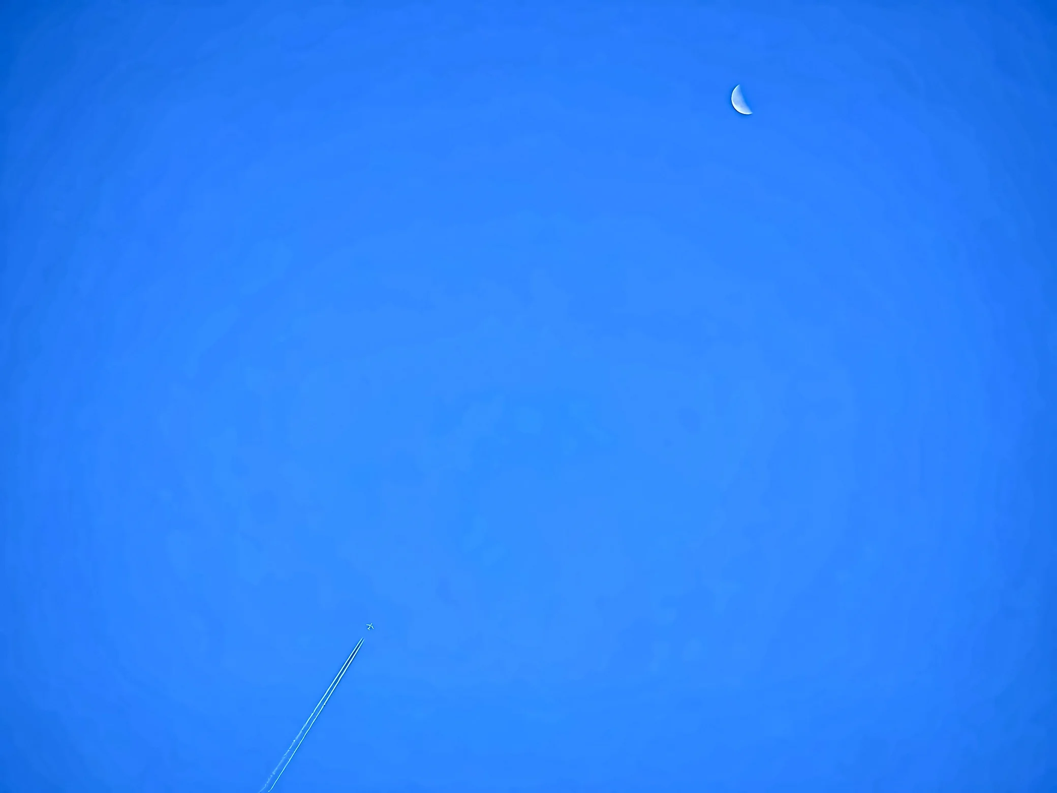 A pale crescent moon in the upper right shares a clear blue sky with a distant jet and twin contrails in the lower left.