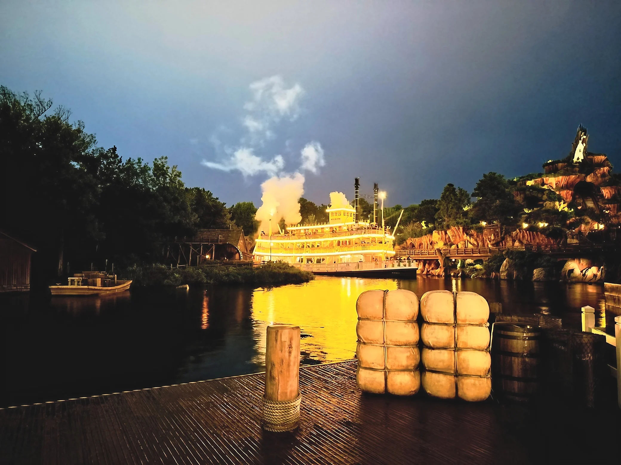 A glowing riverboat and Splash Mountain overlook reflective water behind a wooden dock with bundled props, barrels and a rope-wrapped post.