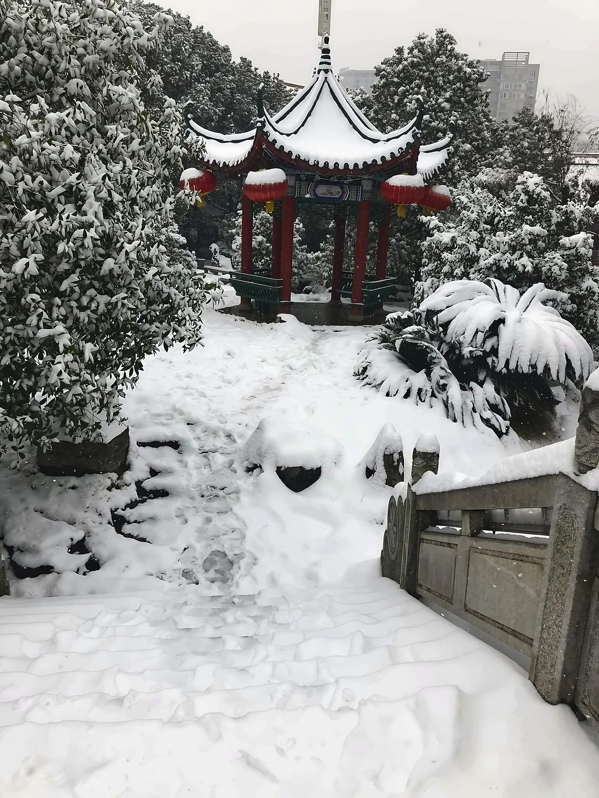 A traditional Chinese pavilion with red lanterns stands among snow-covered trees, stone railings, and steps marked by footprints.