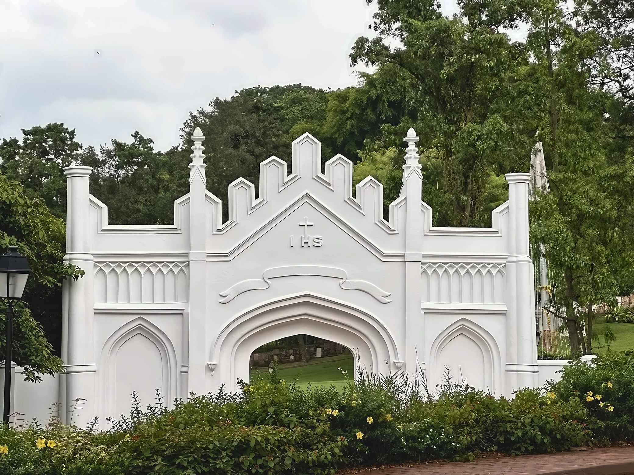 A white Gothic gate bearing a cross and the letters IHS stands among trees and flowers, with a black lamp beside it and lawn visible through the arch.