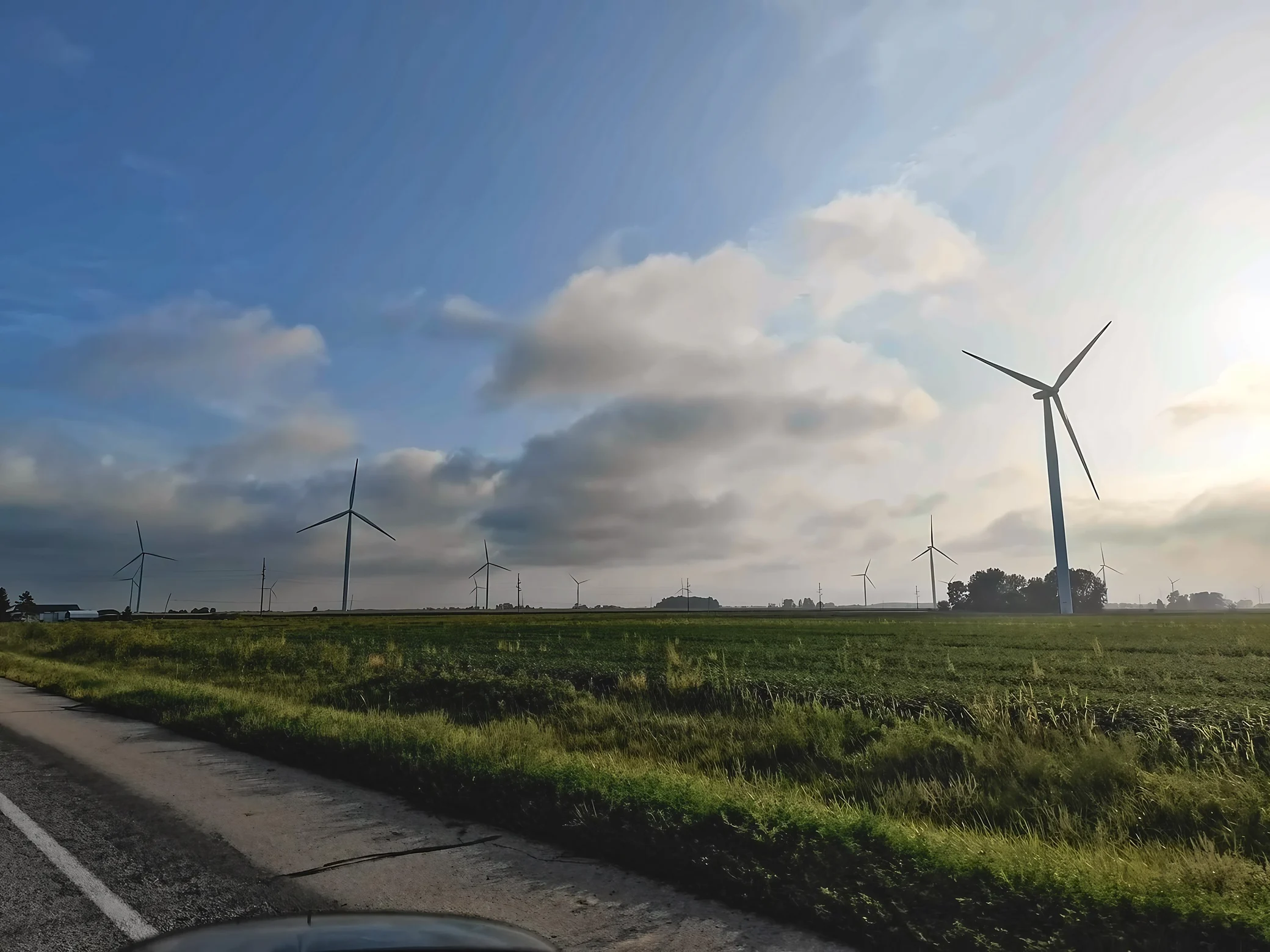 Wind turbines of varying apparent sizes stand across flat farmland beneath a blue sky and layered clouds, with a large turbine on the right.