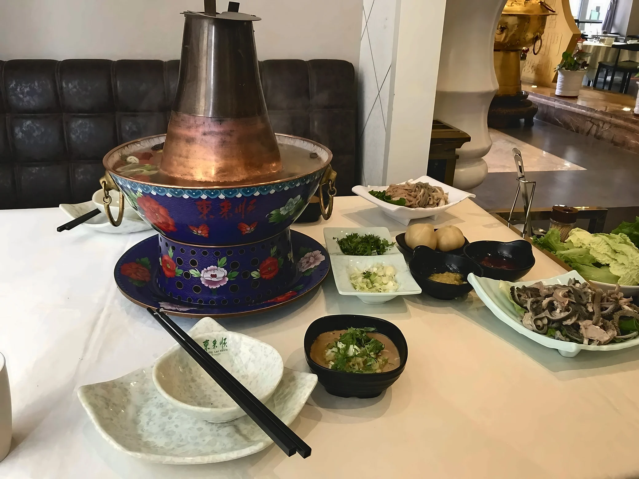 A blue floral copper hotpot bearing the Dong Lai Shun name sits on a restaurant table with tripe, napa cabbage, chopped scallions and dipping sauces.
