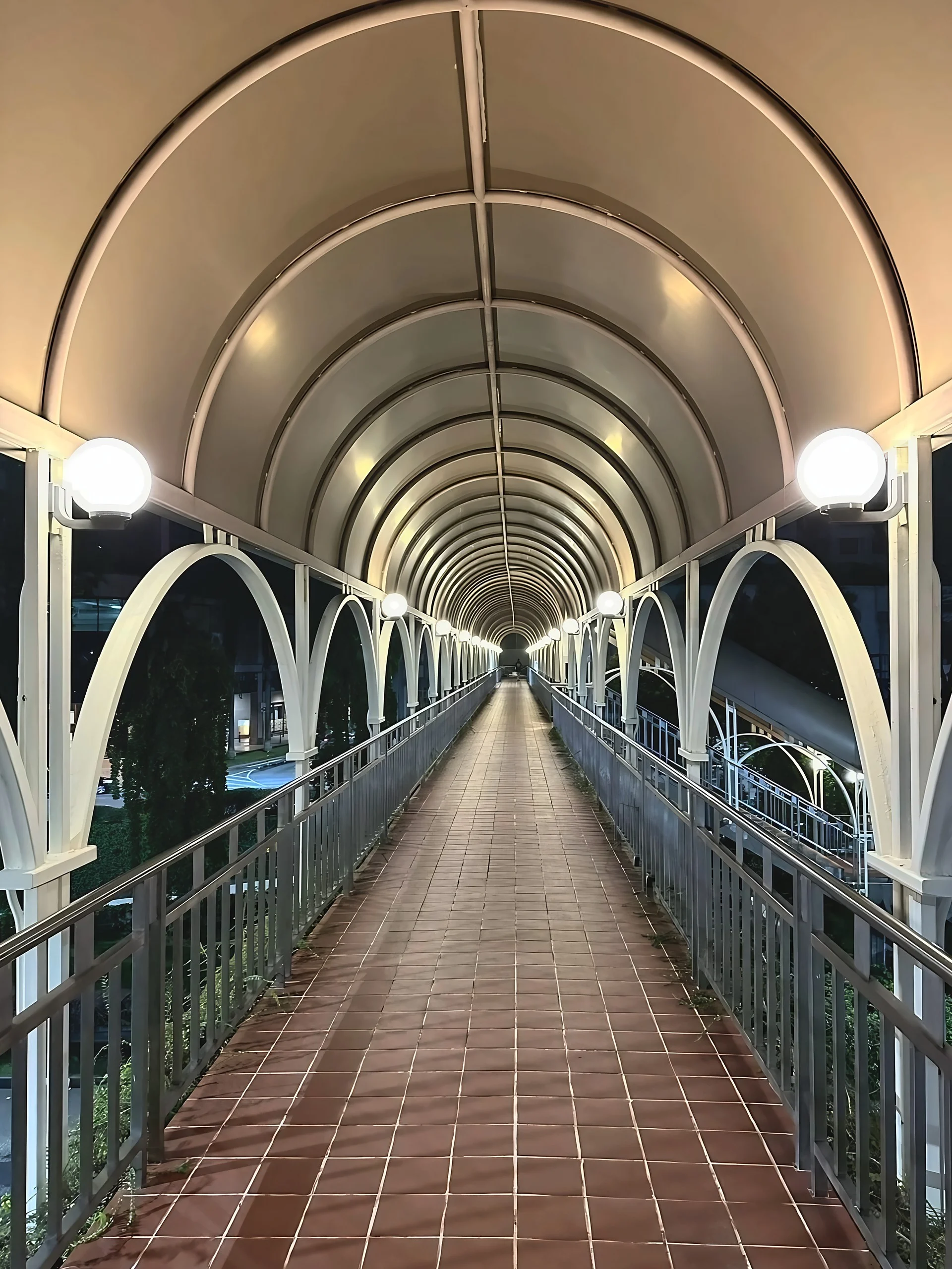 Globe lights illuminate a covered pedestrian bridge where repeating arches, metal railings and reddish tiles converge into the distance.