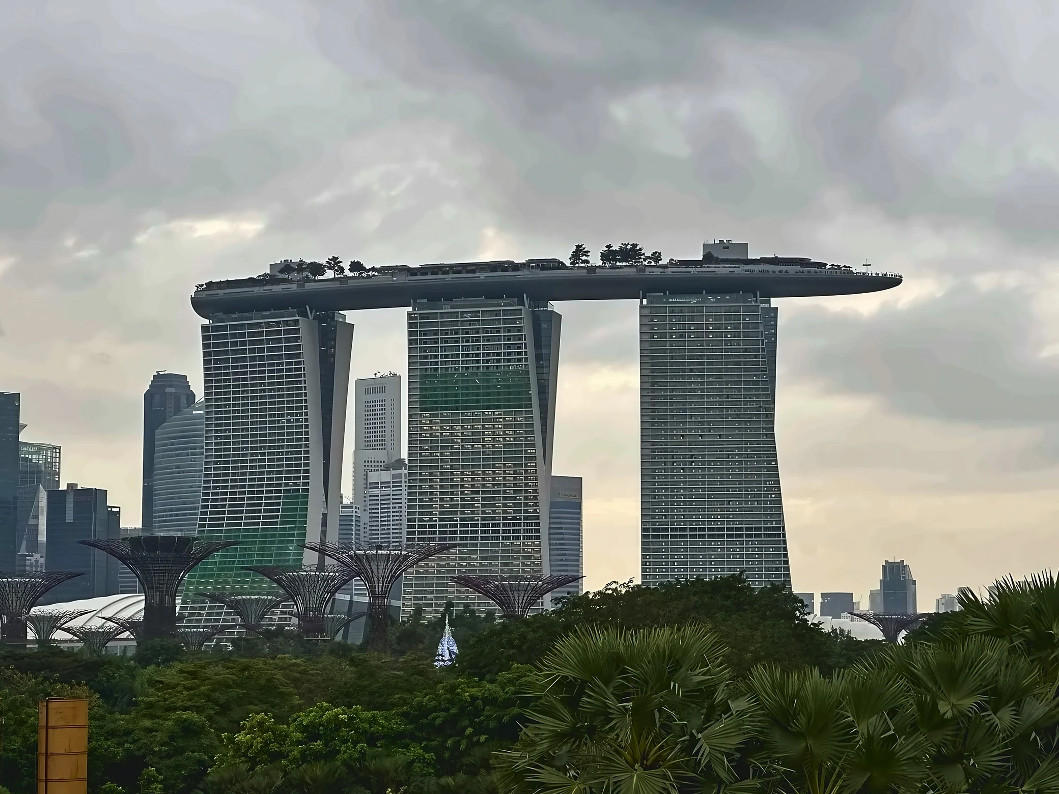Marina Bay Sands towers above the Supertrees and dense tropical foliage beneath an overcast sky.
