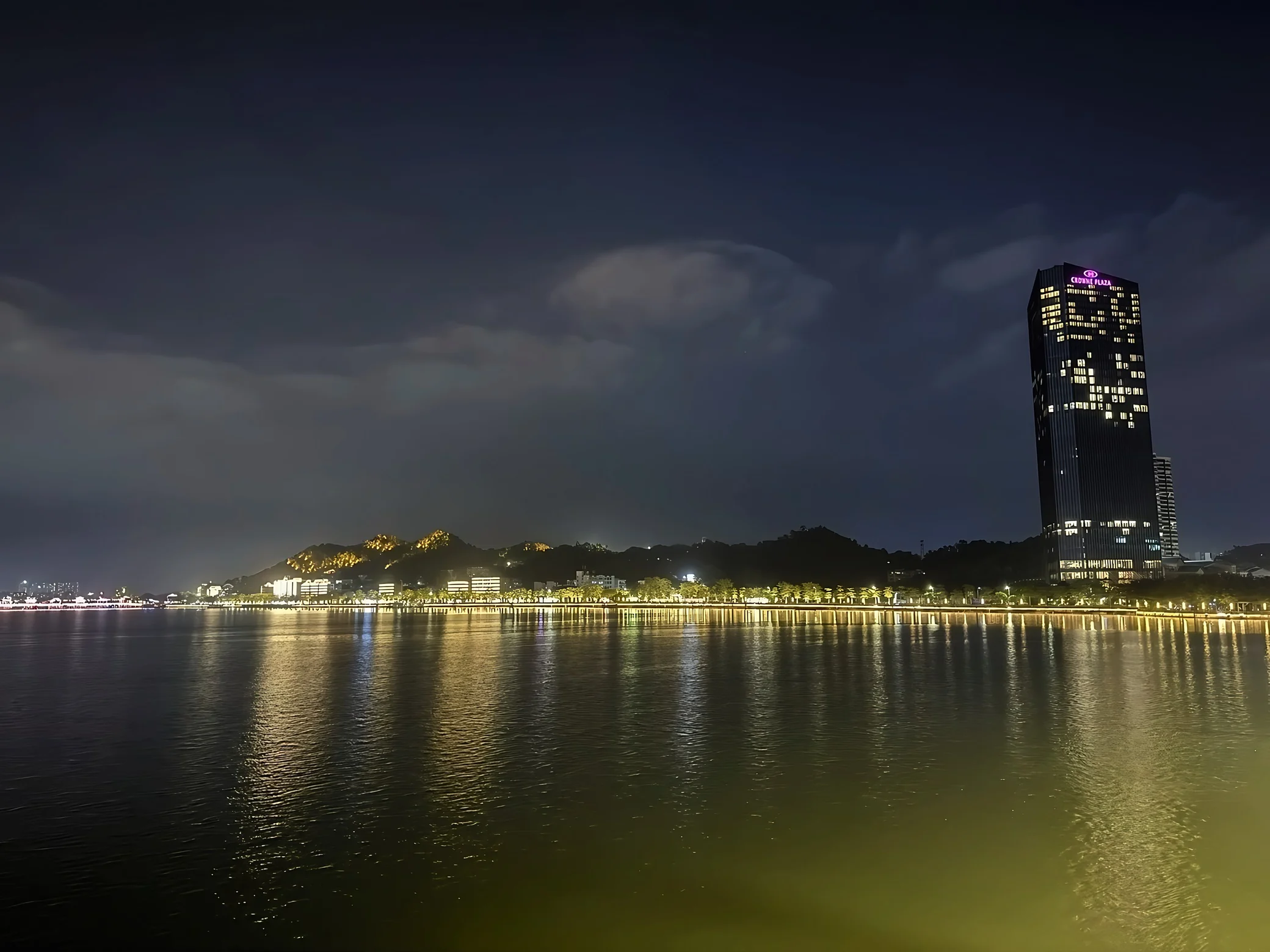 Waterfront lights and a tall illuminated building reflect across the river beneath a clouded sky and distant hills.