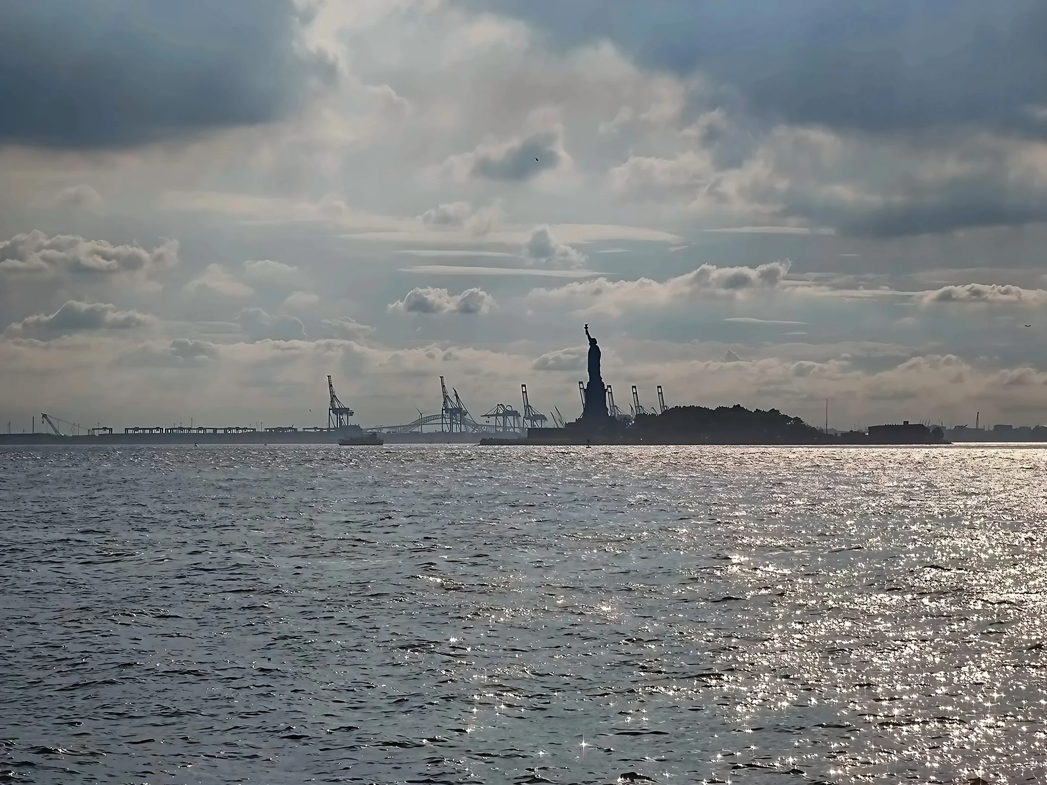 The Statue of Liberty appears in silhouette across shimmering harbor water, with port cranes and layered clouds behind it.