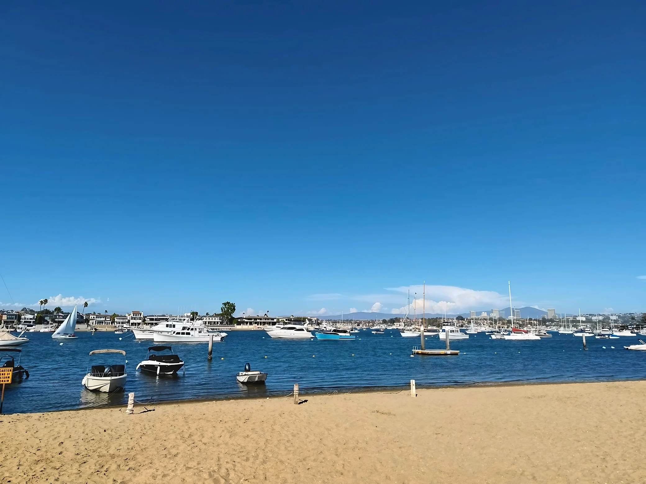 Yachts, sailboats and small boats float in a blue harbor beside a sandy beach, with waterfront buildings across the water.