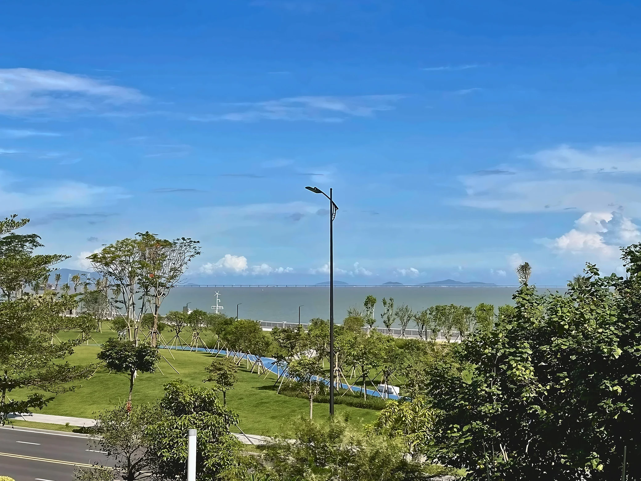 A landscaped waterfront park in Zhuhai overlooks the sea, a long bridge and distant hills beneath a broad blue sky.