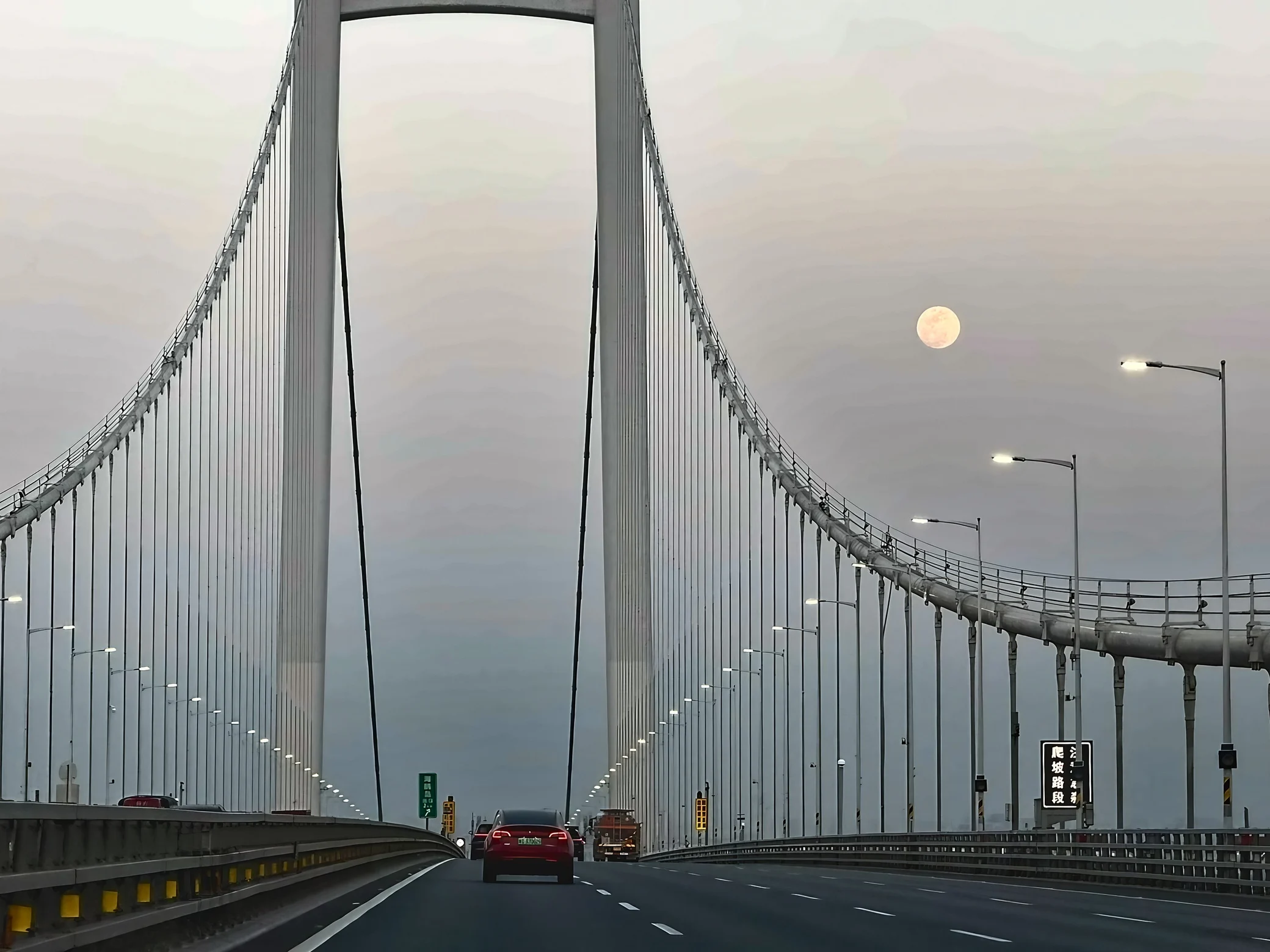 A pale moon hangs beside a white suspension bridge, with a red car and trucks ahead beneath illuminated streetlights.