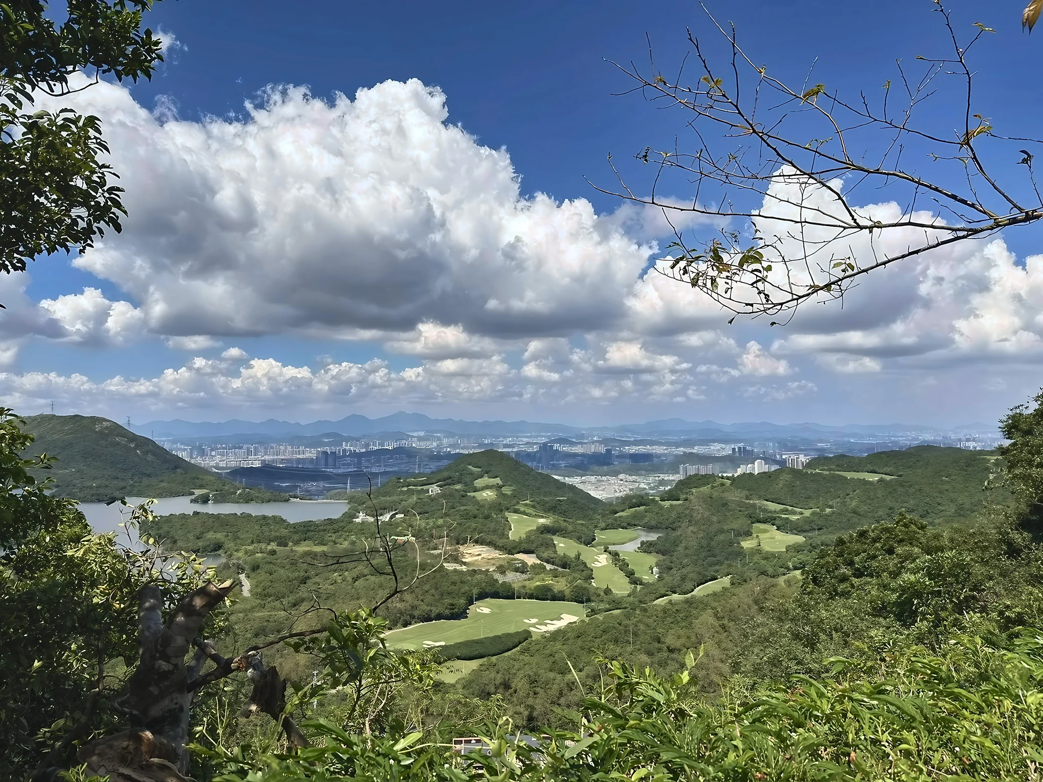 Large white clouds rise above a wooded valley with golf fairways, lakes, and a distant city framed by nearby foliage.