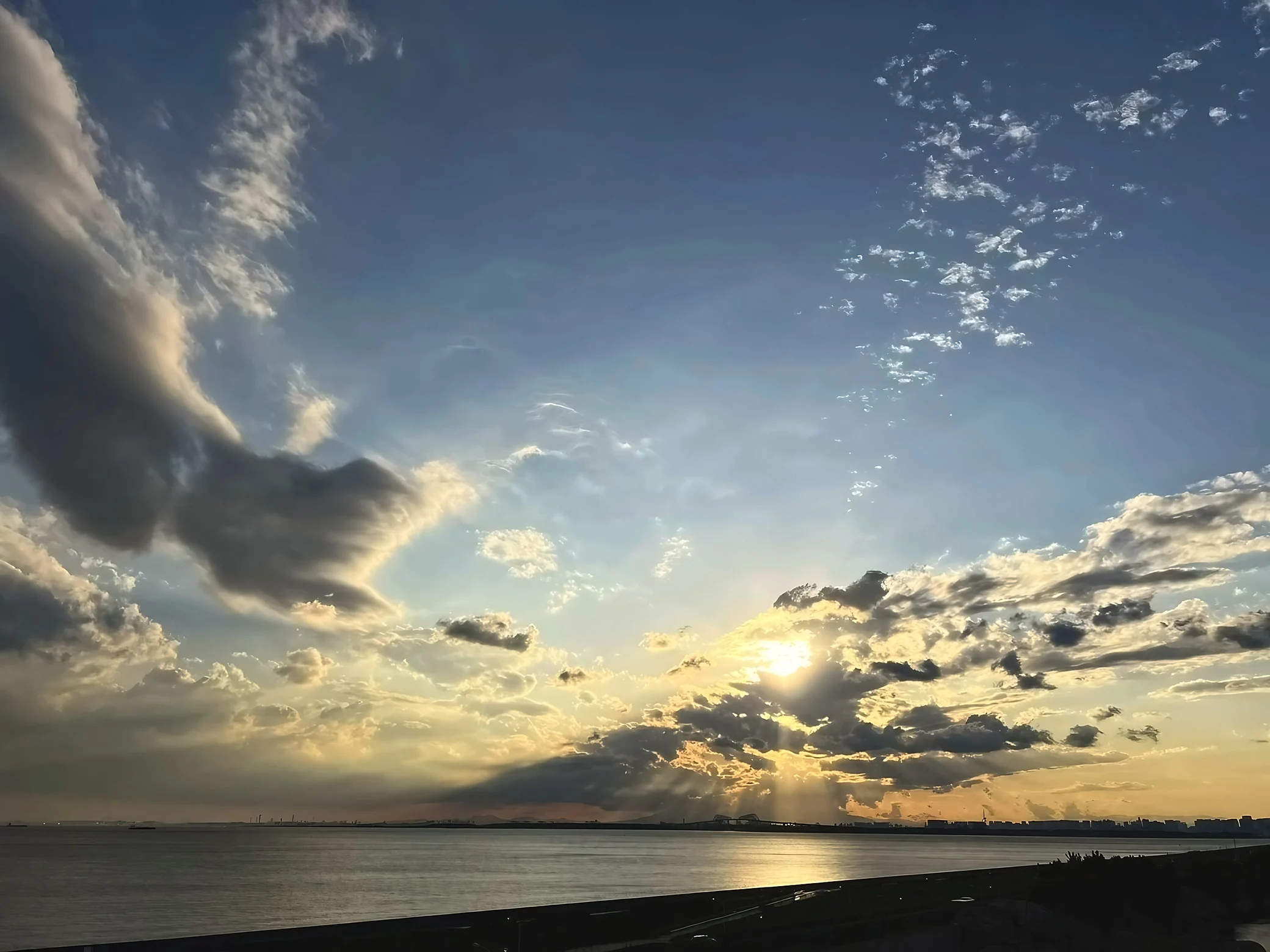 Golden rays fan out through thick clouds over calm bay waters and a silhouetted distant shoreline.