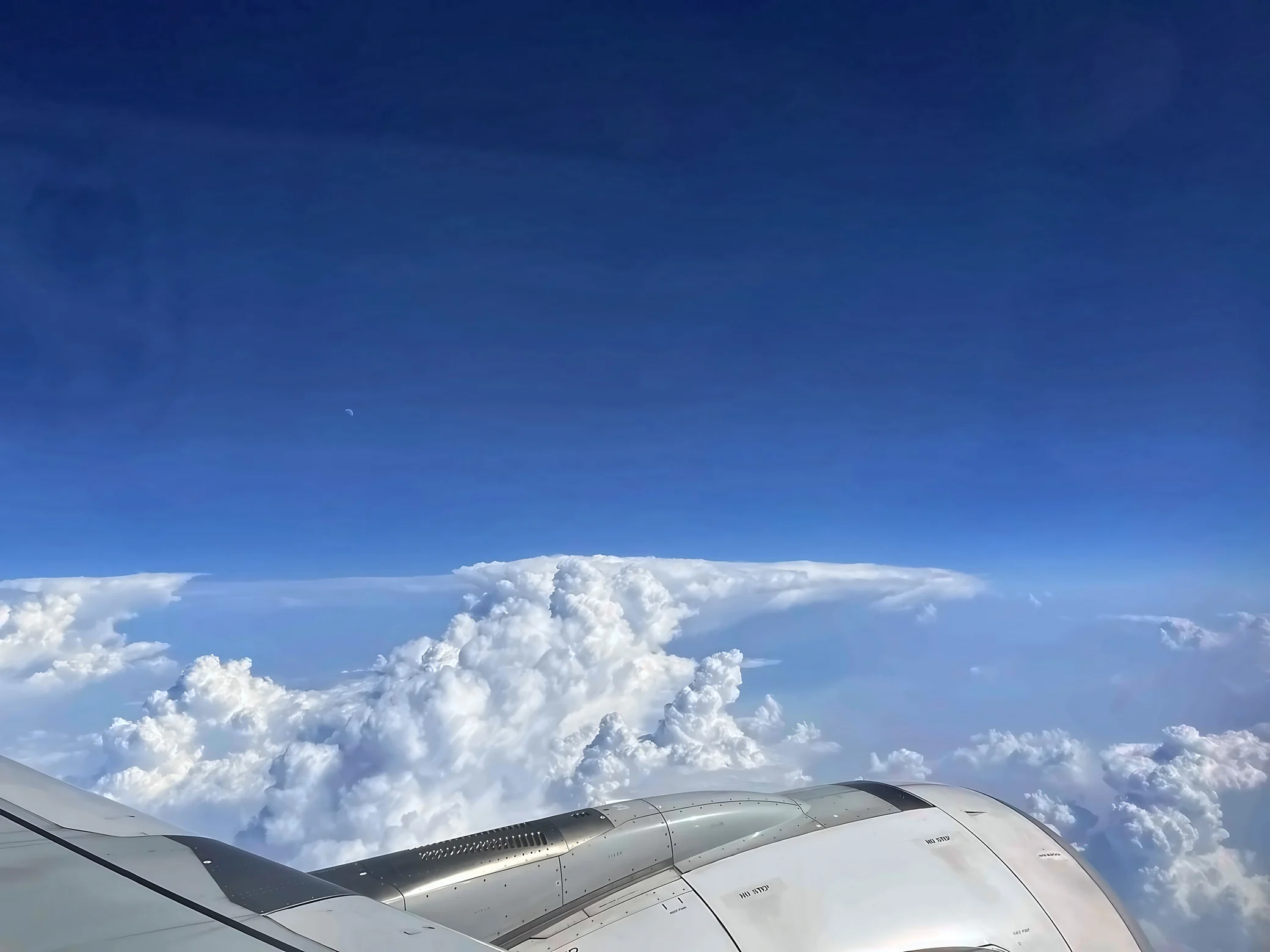 An airplane wing and engine fill the foreground beneath white cloud towers, blue sky, and a faint moon.
