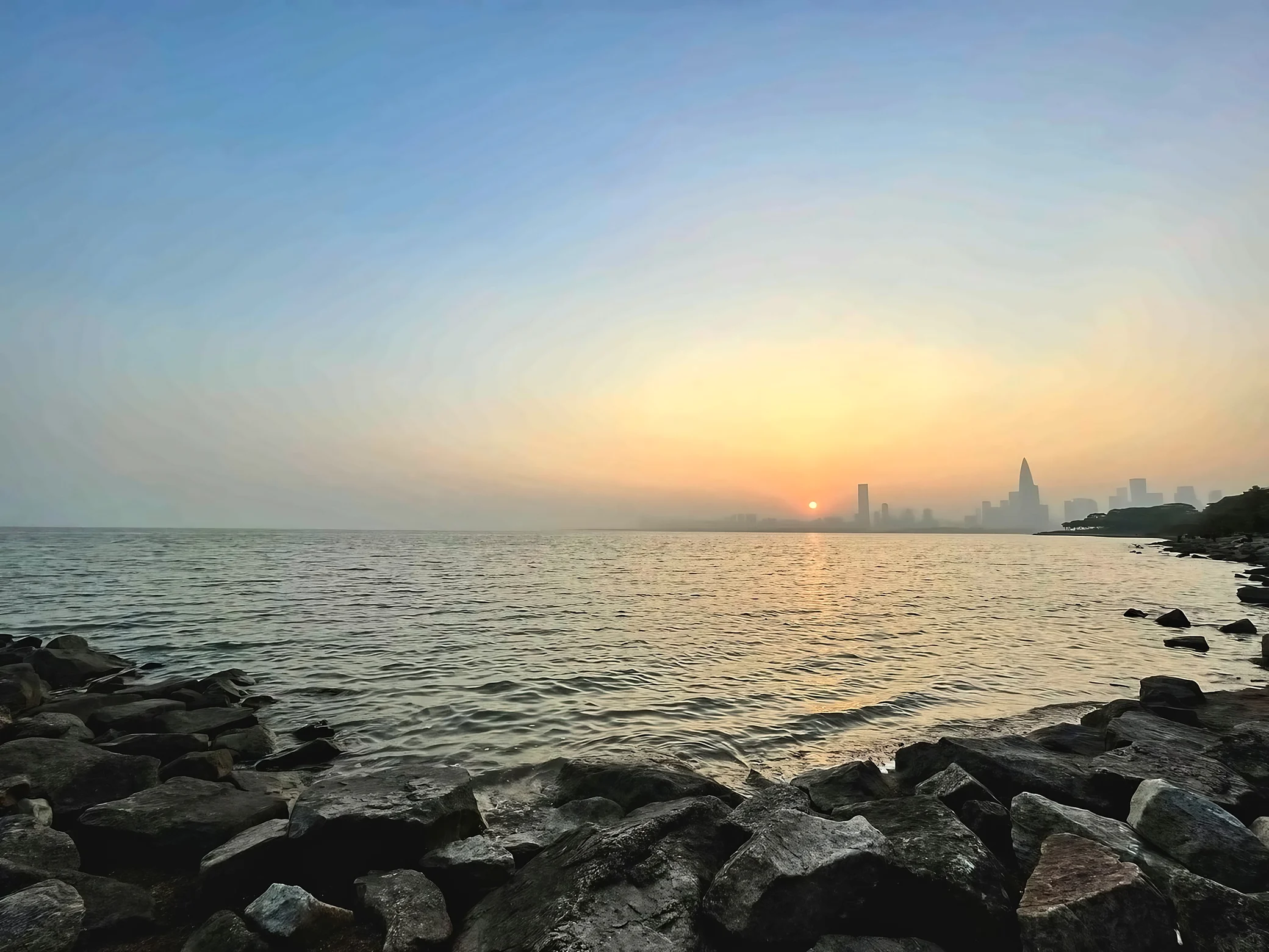 Dark shoreline rocks frame a broad bay with a golden sun reflection and a hazy city skyline stretching along the distant shore.