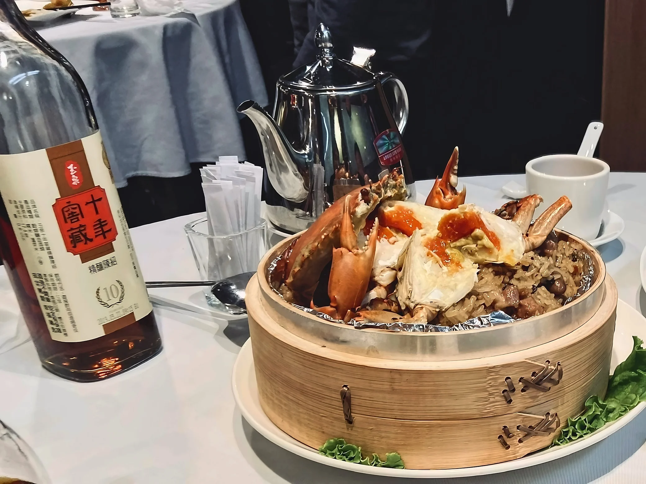 Crab pieces and orange roe top sticky rice in a bamboo steamer, beside a bottle of aged Yu Chuan wine, a metal teapot, and a white teacup.