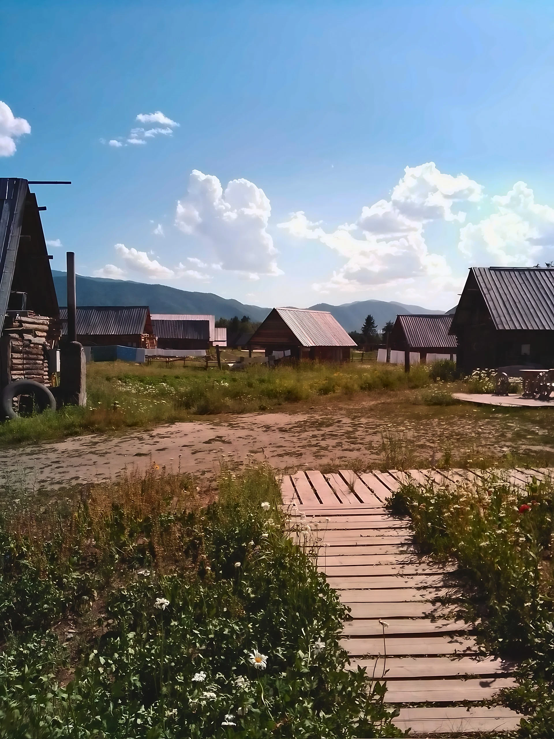 A wooden path crosses flowering grass toward scattered pitched-roof log cabins, with mountain ridges beneath a blue sky filled with white clouds.