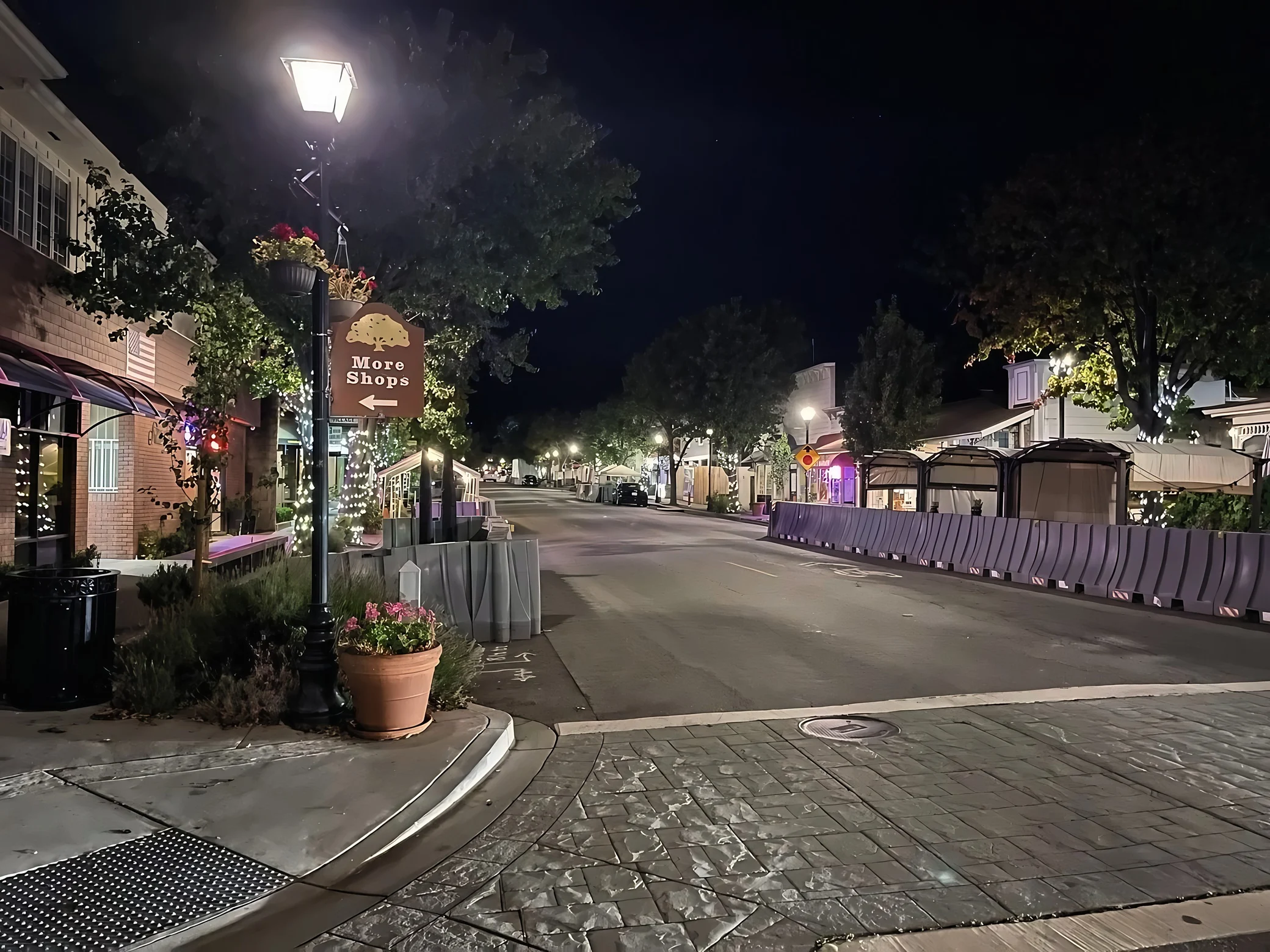 A small-town California street is lined with low-rise shops, illuminated trees and covered outdoor seating beside a “More Shops” sign pointing left.
