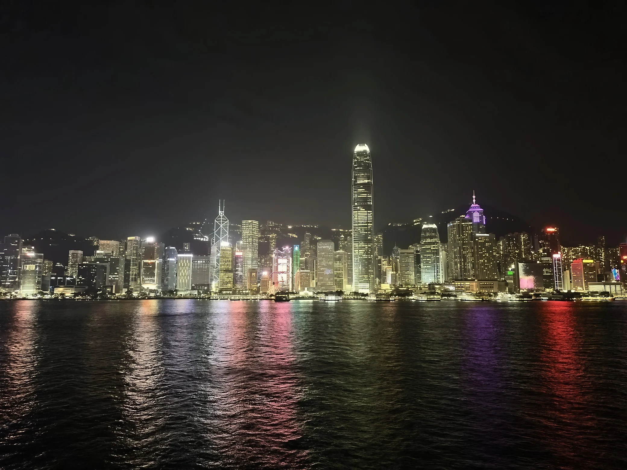 Hong Kong Island's illuminated skyline stretches across Victoria Harbour, with Two IFC, the Bank of China Tower and the observation wheel above colorful reflections.