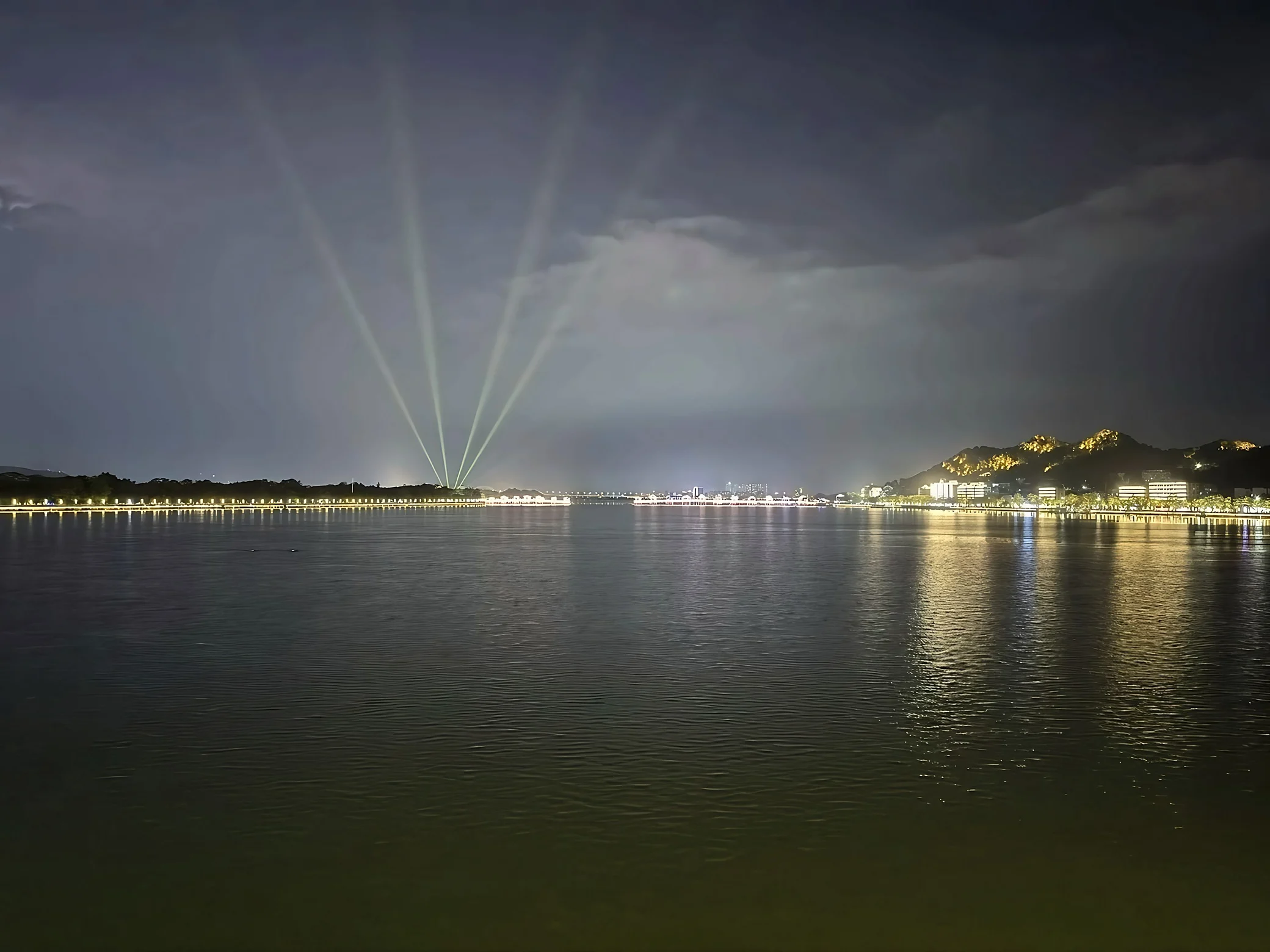 Fan-shaped beams rise above the river as waterfront lights and golden hill outlines reflect across the water.