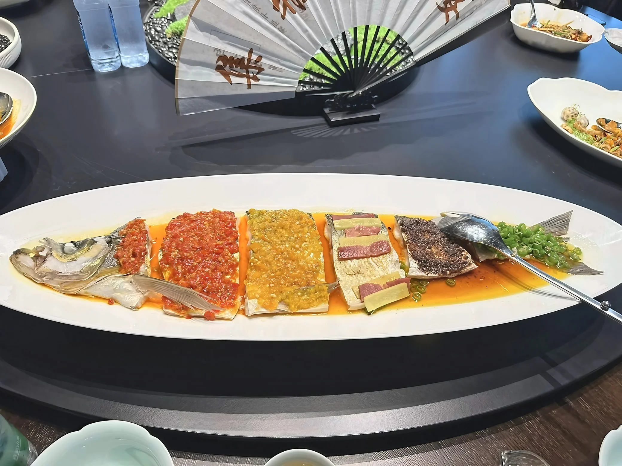 A long white platter holds fish sections topped with red and yellow chopped seasonings, ginger, sliced meat, and scallions, with a calligraphy fan behind it.