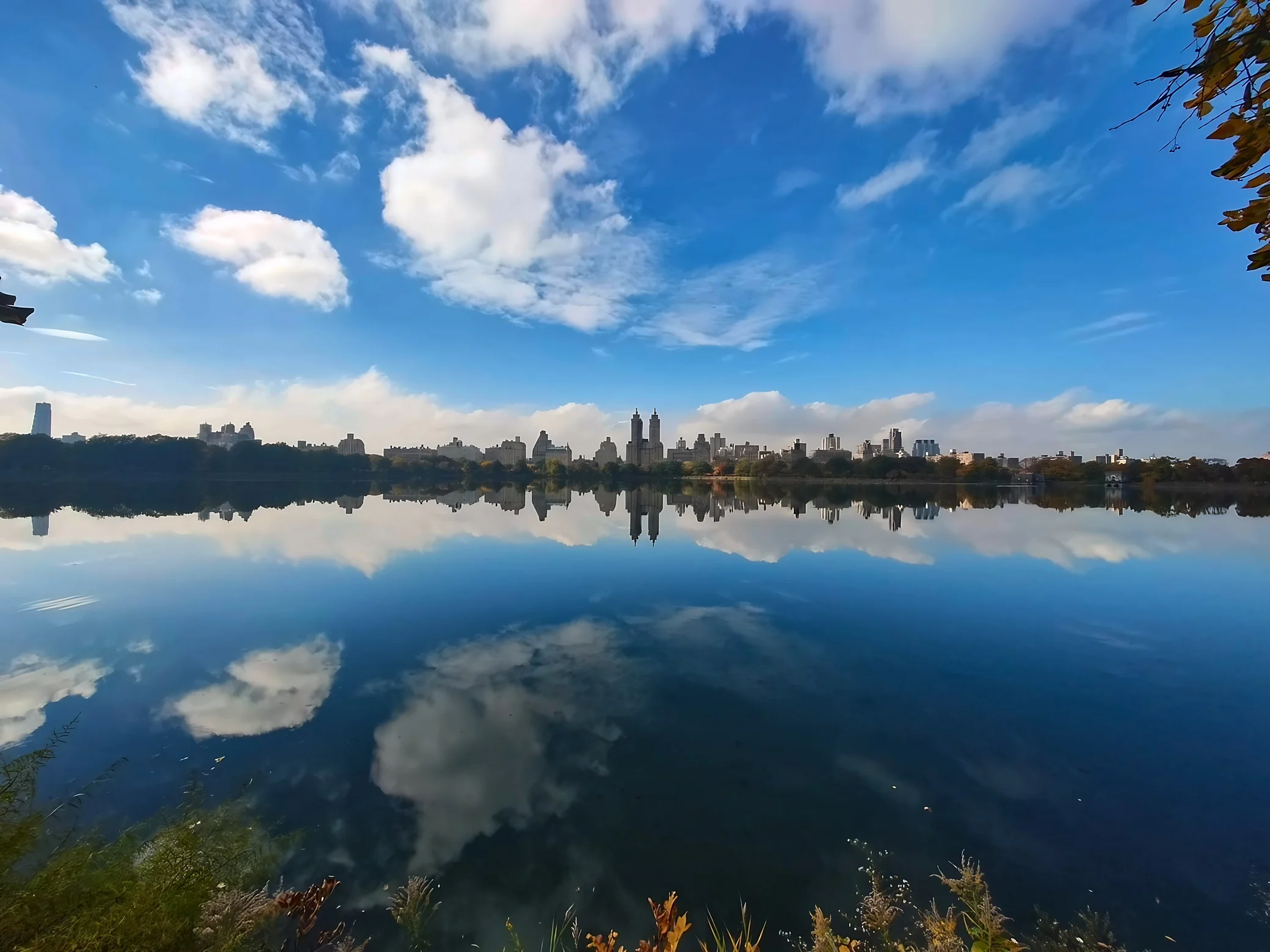 Blue sky, white clouds, and a distant Manhattan skyline reflect across the calm expanse of Central Park's reservoir.