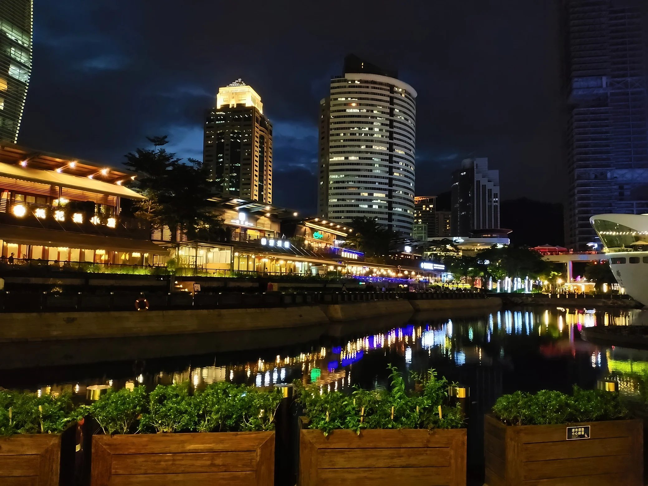 Warmly illuminated waterfront restaurants and high-rise buildings reflect in the water, with a white ship bow at the right and planters in front.