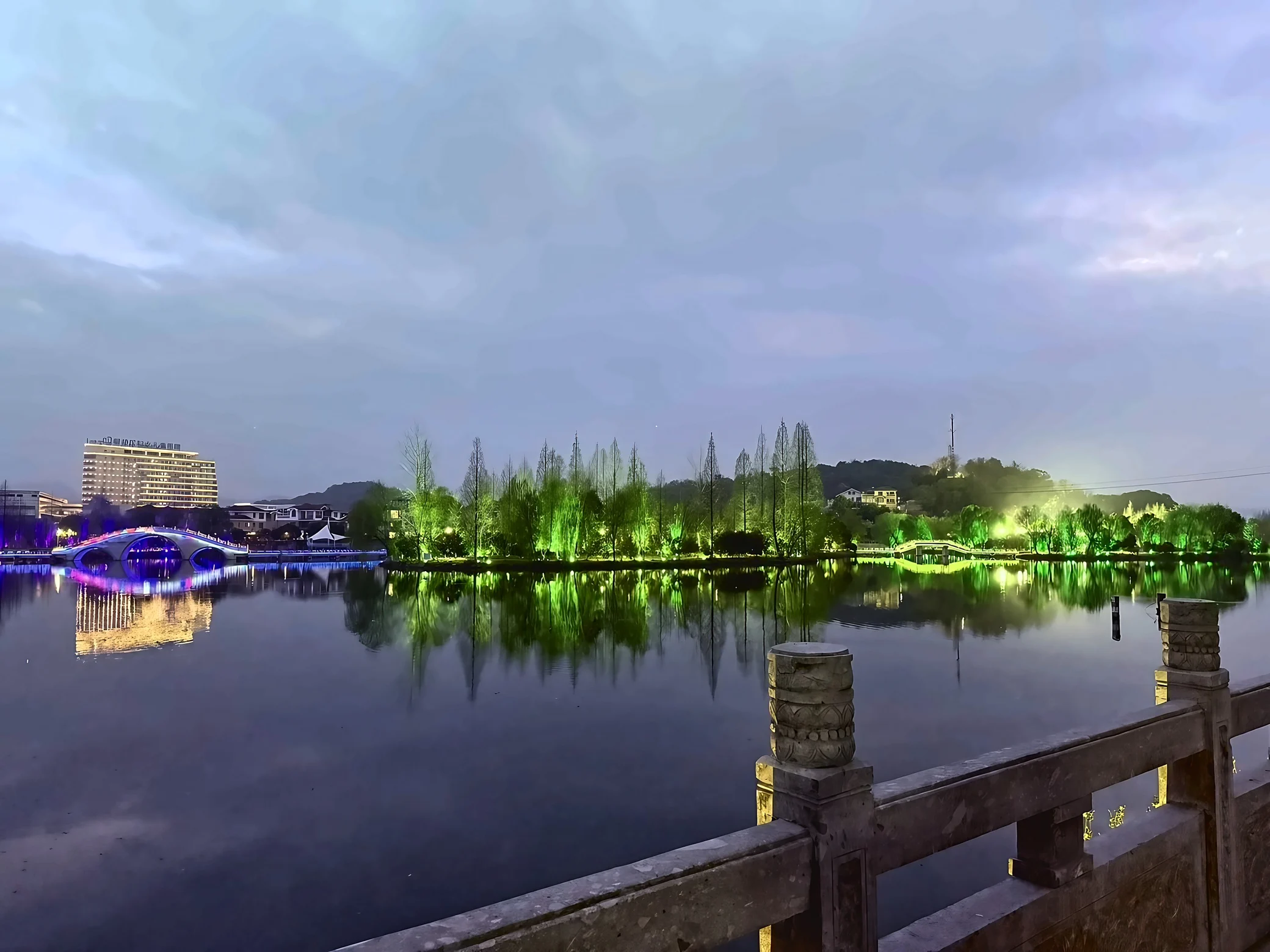 Green-lit trees, a purple-blue arch bridge, and a golden-lit building reflect in Jianhu beyond a carved stone railing.