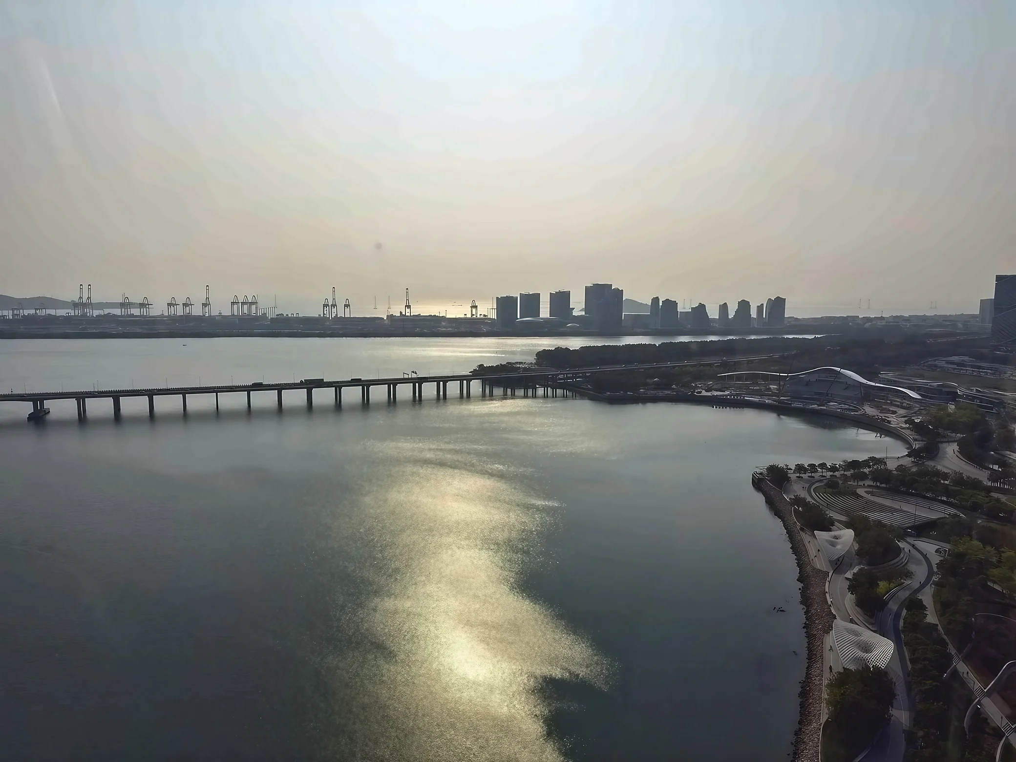 An elevated view of OH Bay shows sunlight shimmering on the water, a long bridge, curved waterfront paths, white shade structures and distant port cranes and towers.