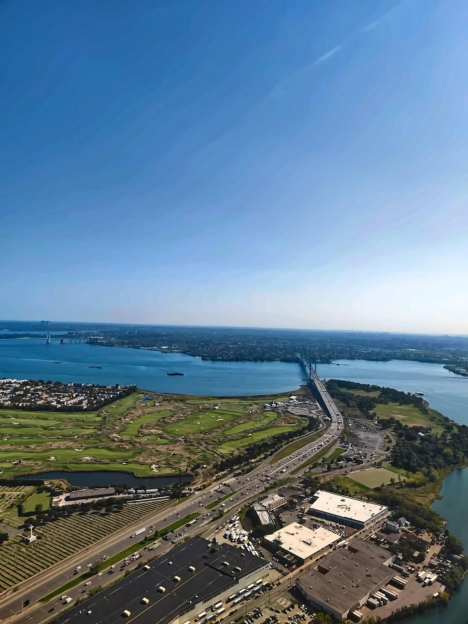 An aerial view follows a multilane highway toward the Bronx–Whitestone Bridge, passing a golf course, cemetery and waterfront industrial buildings.