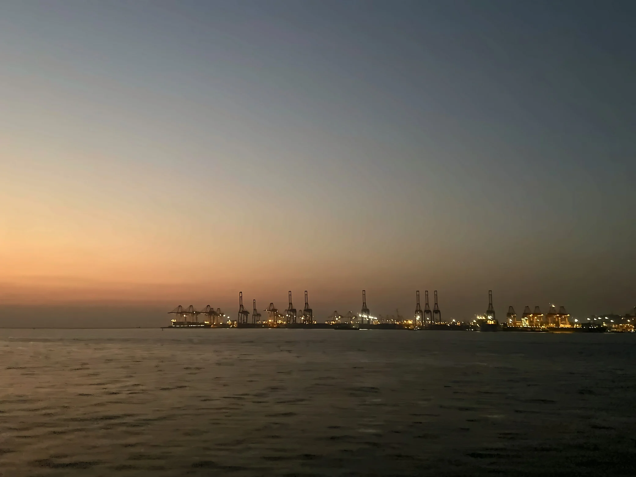An orange sky hangs over open water, with illuminated docks and rows of cranes along the right side.